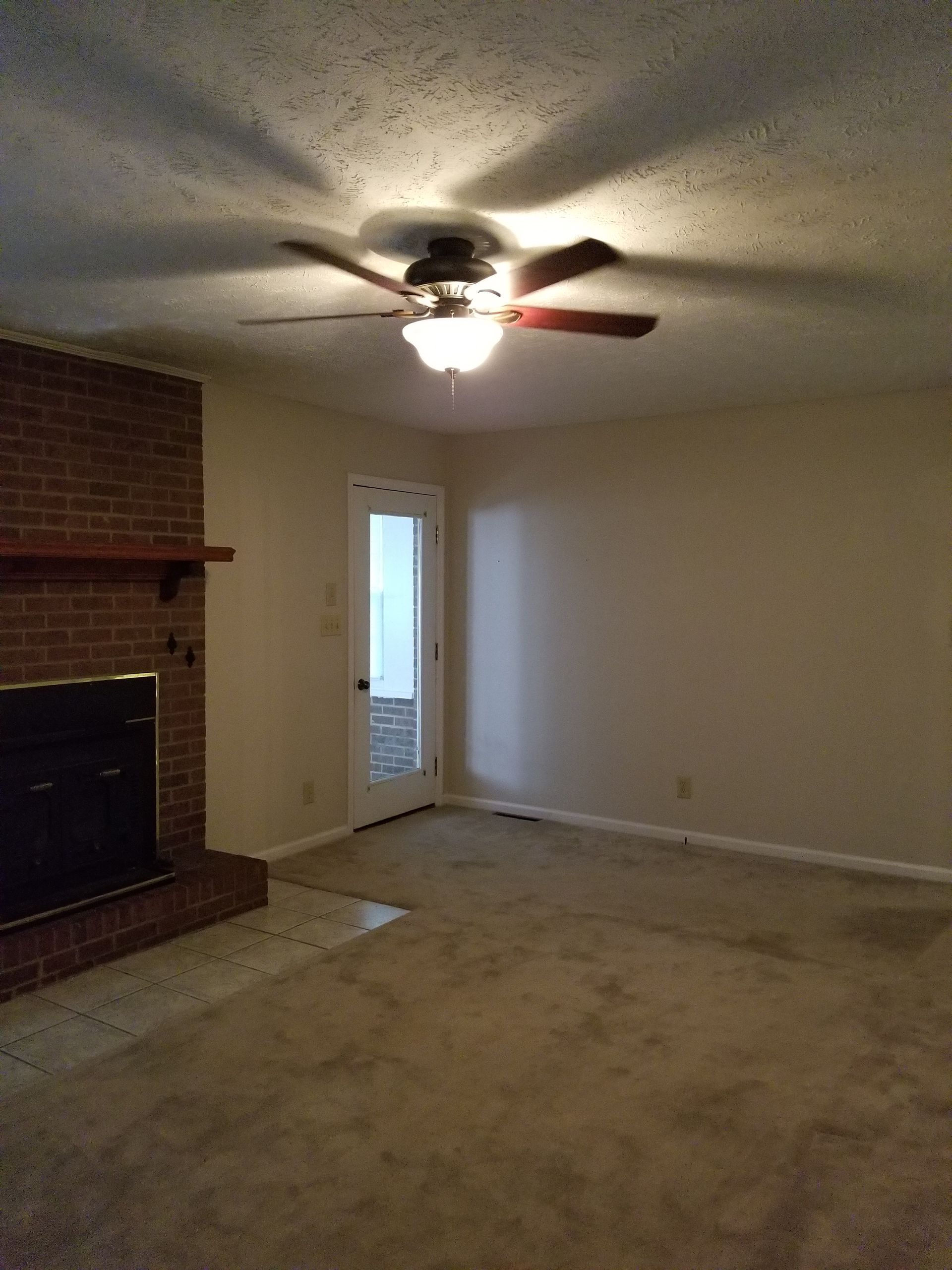 Empty living room with brick fireplace, beige carpet, and a ceiling fan with lights on.