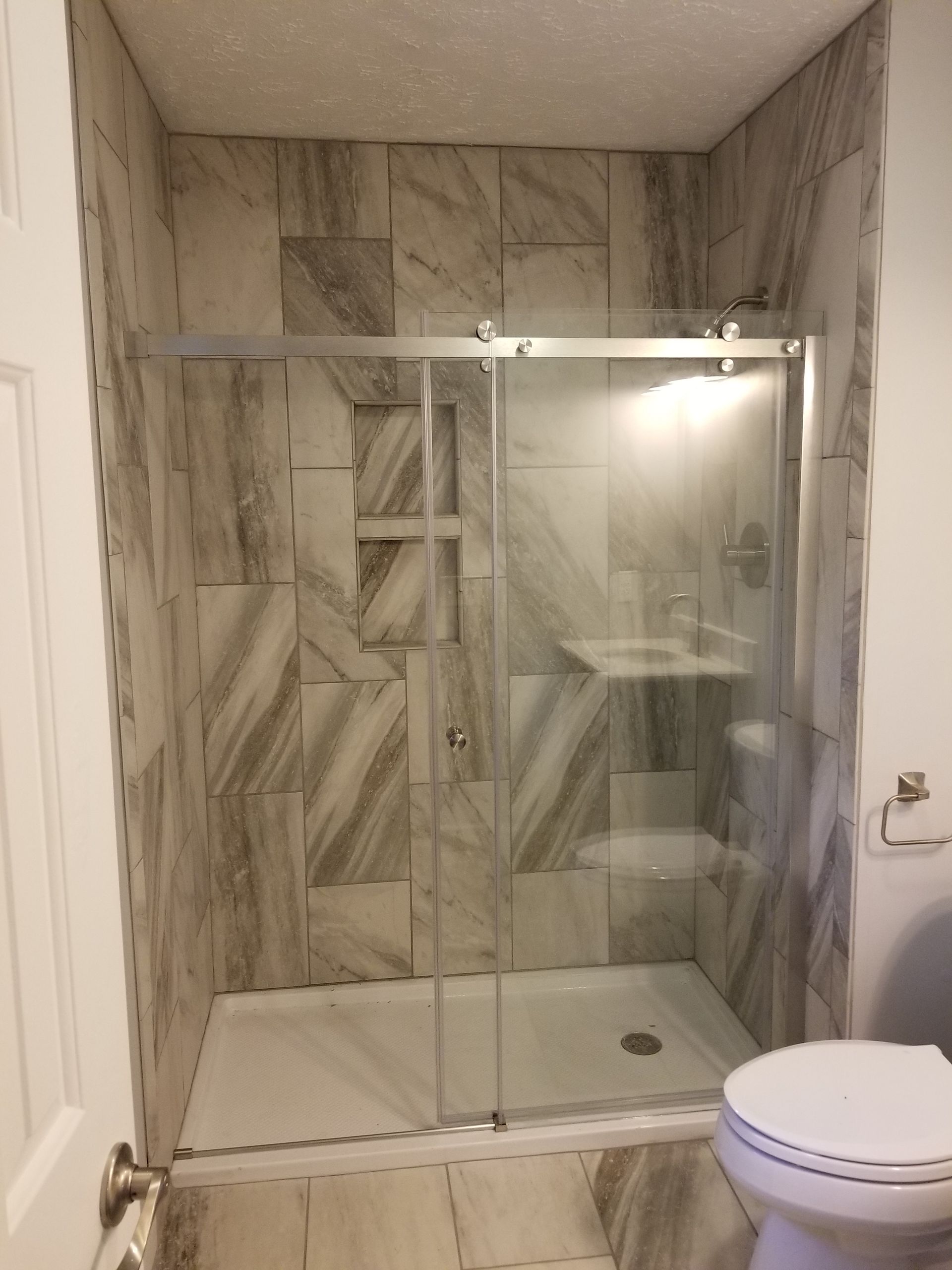 Walk-in shower with glass sliding doors and gray tile walls, next to a toilet.