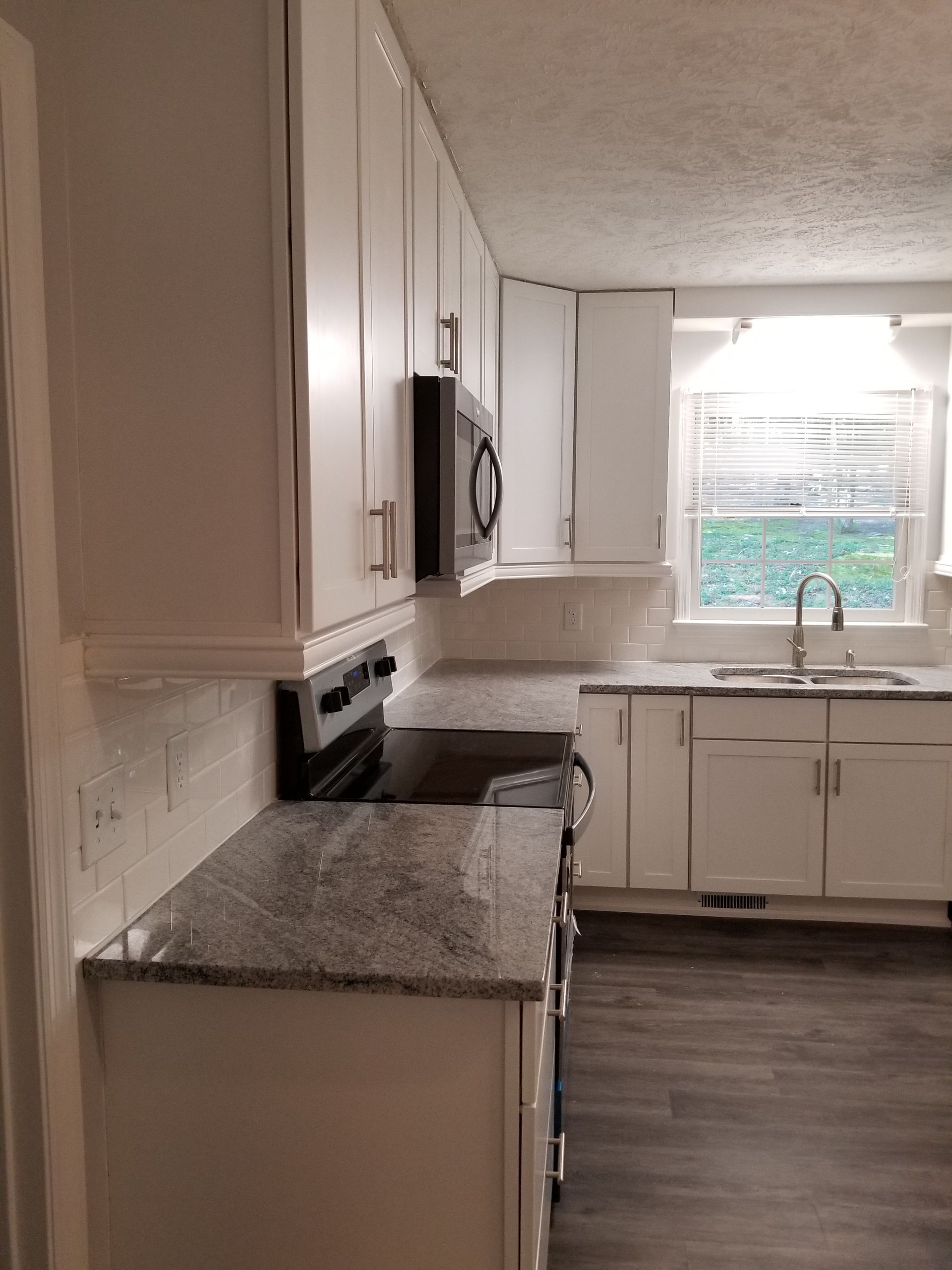 Small kitchen with white cabinets, granite counters, dark wood floor, and a bright window over the sink.