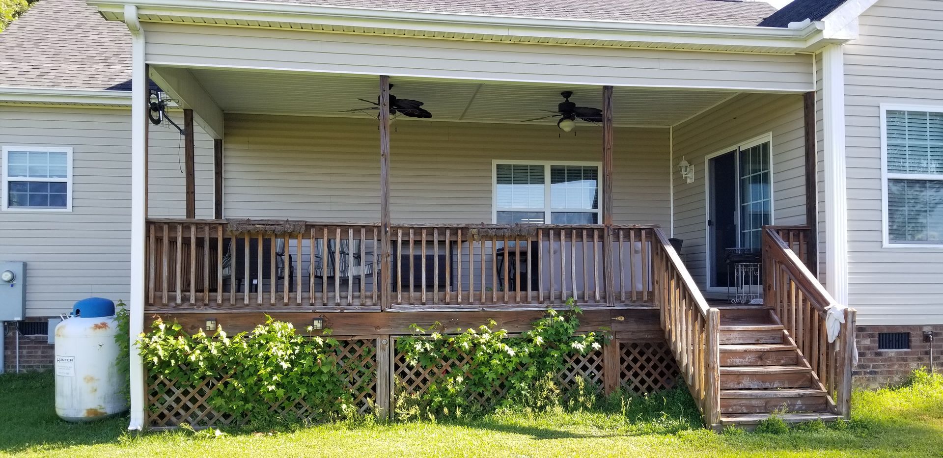 Covered front porch with wooden railing and steps on a beige house, with a white propane tank on the lawn.