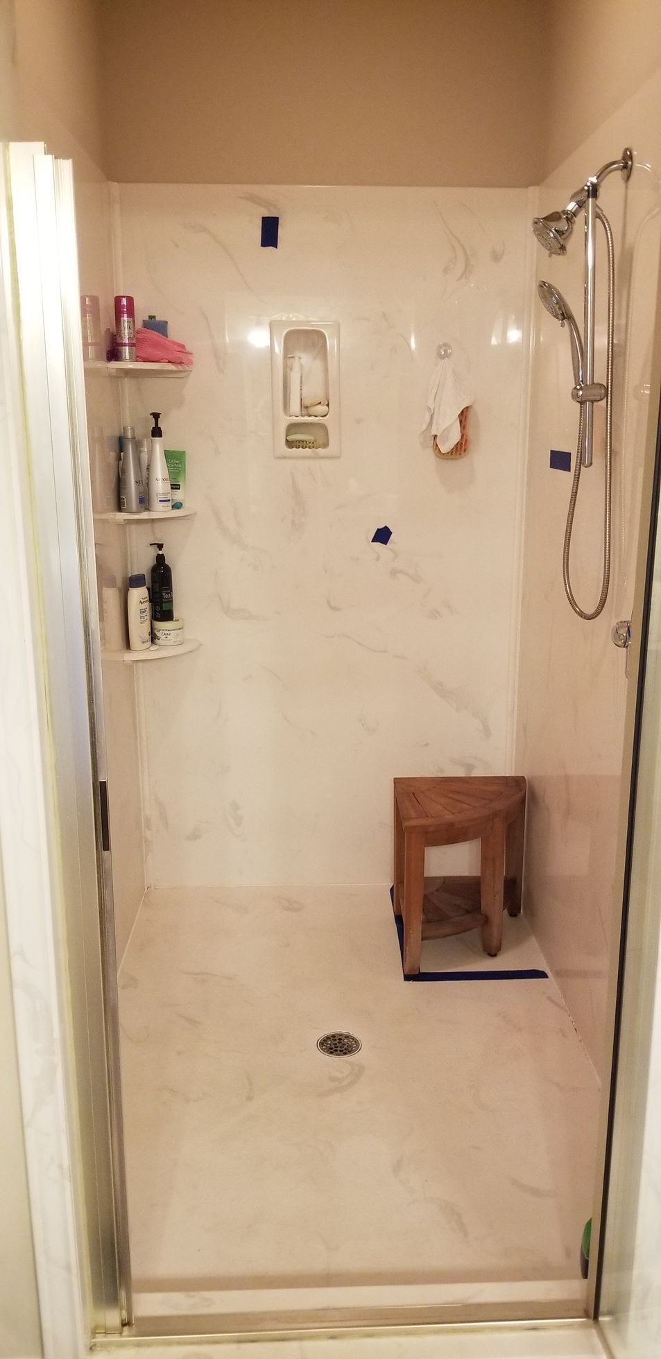 Empty tiled shower stall with glass door, built-in shelf, handheld showerhead, and a small wooden stool inside