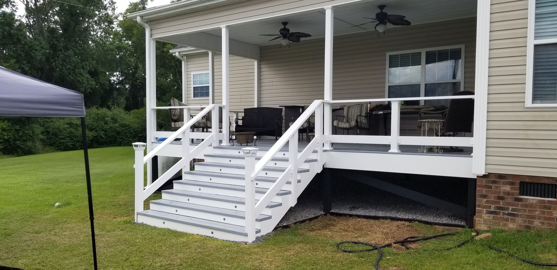 White wooden porch ramp and stairs attached to a beige house with black ceiling fans and outdoor seating.