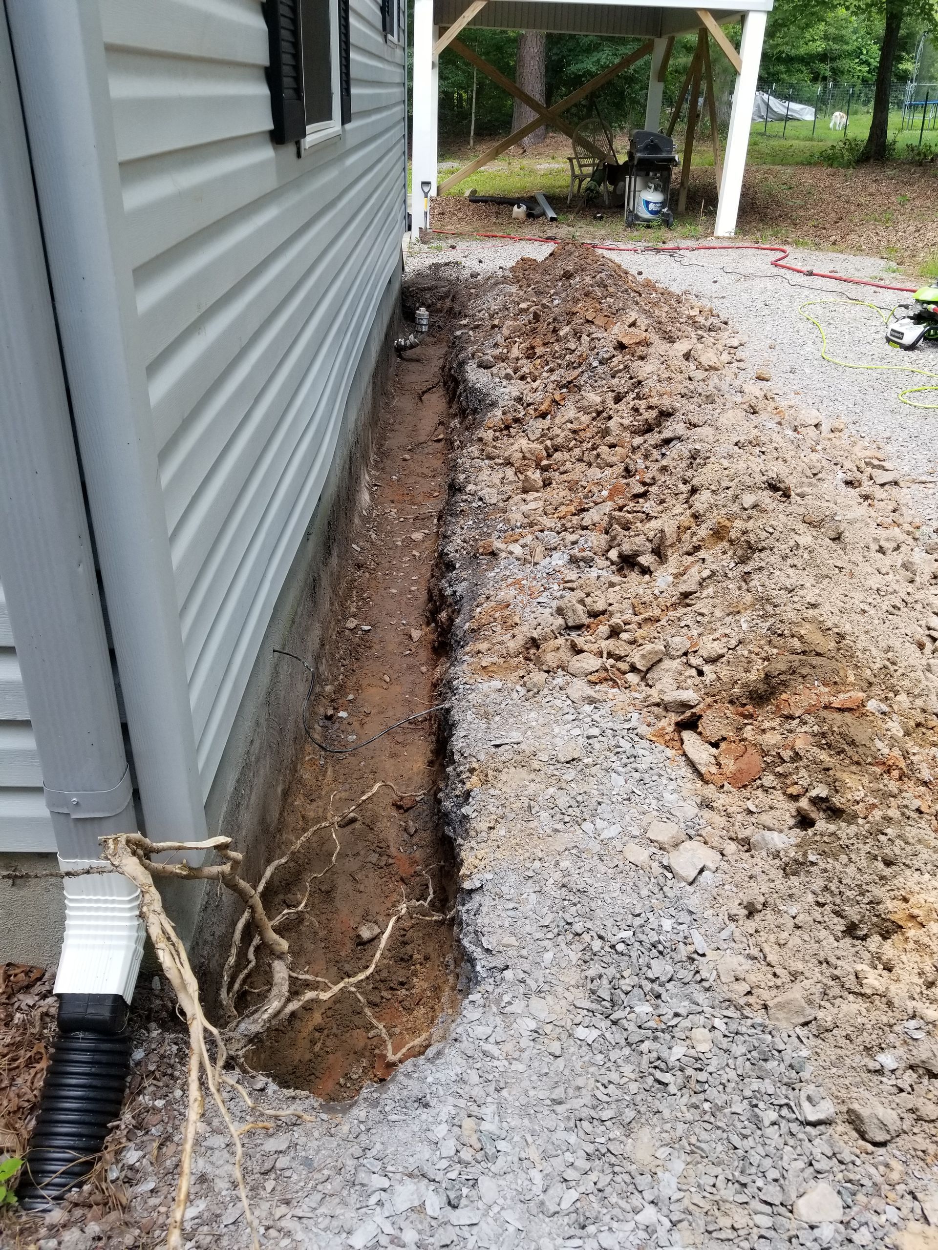 Trench dug beside gray house siding, with soil piled along the gravel driveway.