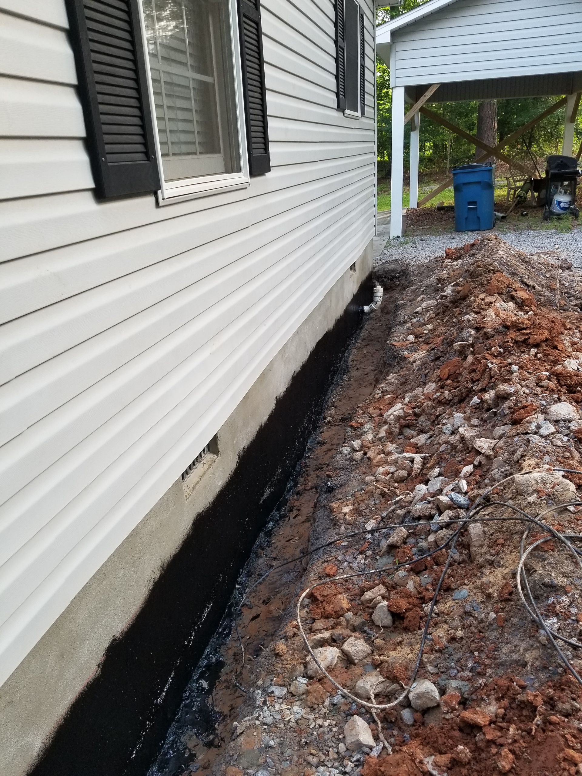 Side of white house with exposed foundation next to a narrow trench of dirt and gravel.