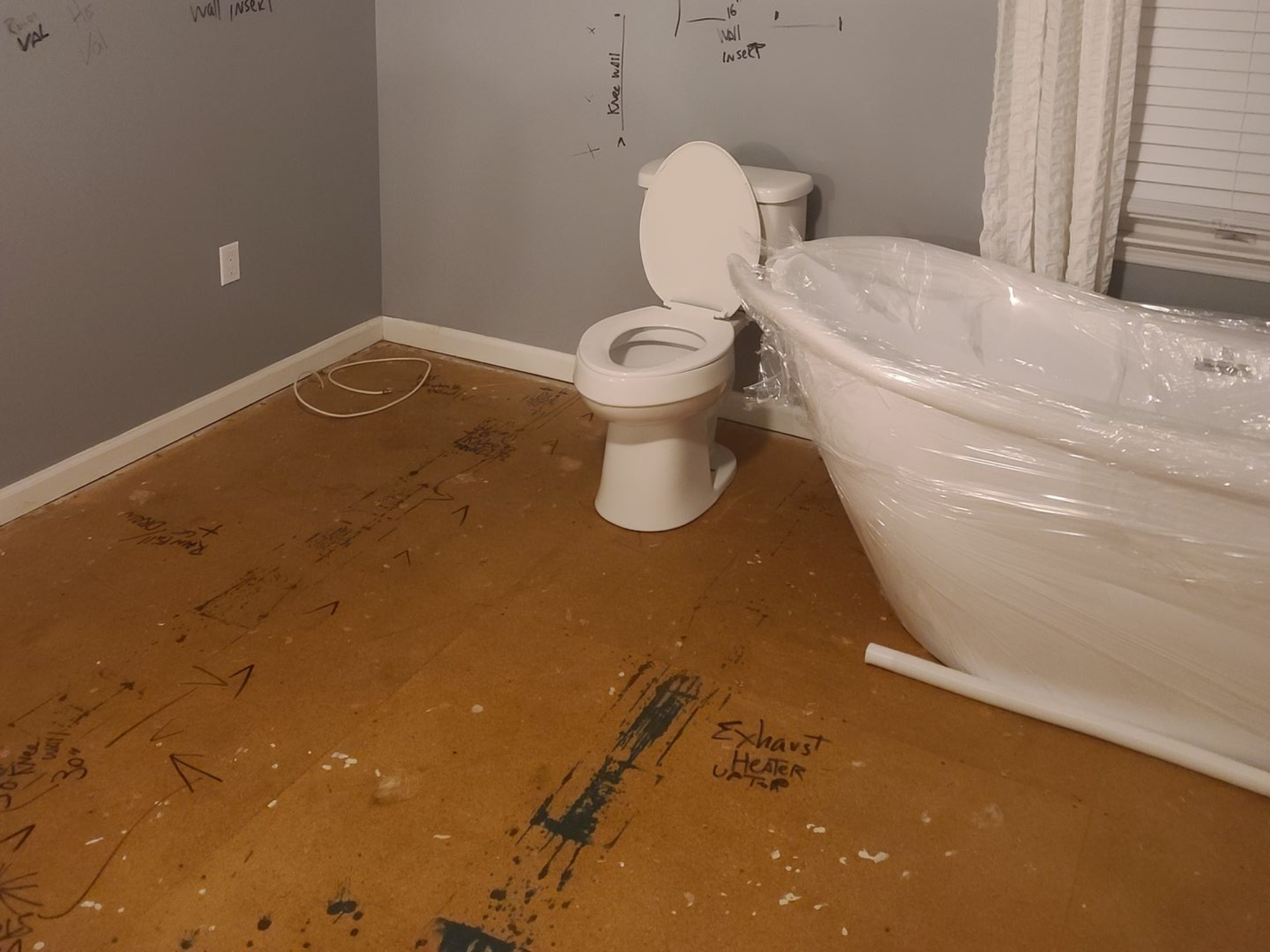 Bathroom under renovation with bare plywood subfloor, toilet, and a bathtub wrapped in plastic.
