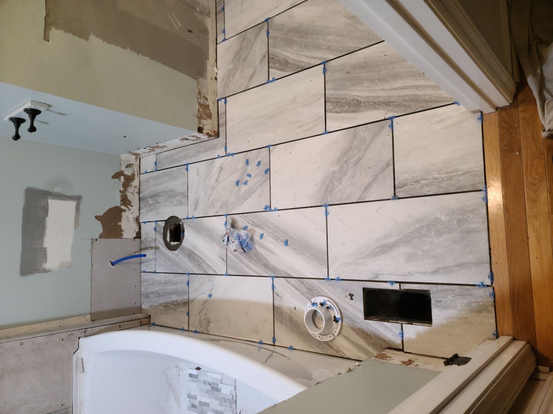 Partially tiled bathroom wall with a tub, plumbing openings, and unfinished drywall above