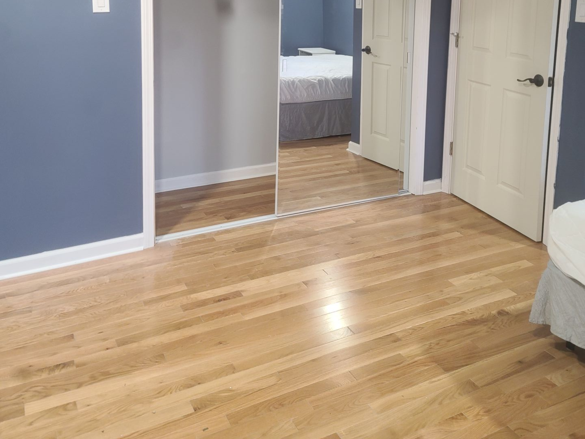 Empty hardwood bedroom with blue walls, open doorways, and a bed visible in the back room.