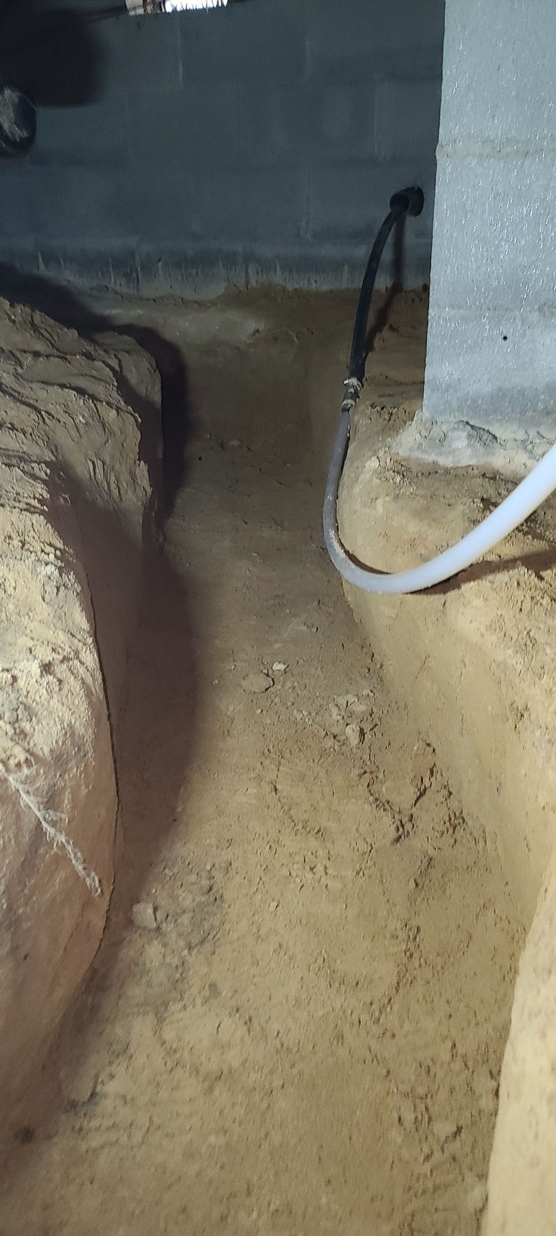 Dimly lit crawl space with dirt floor, concrete wall, and a white cable running along the right side