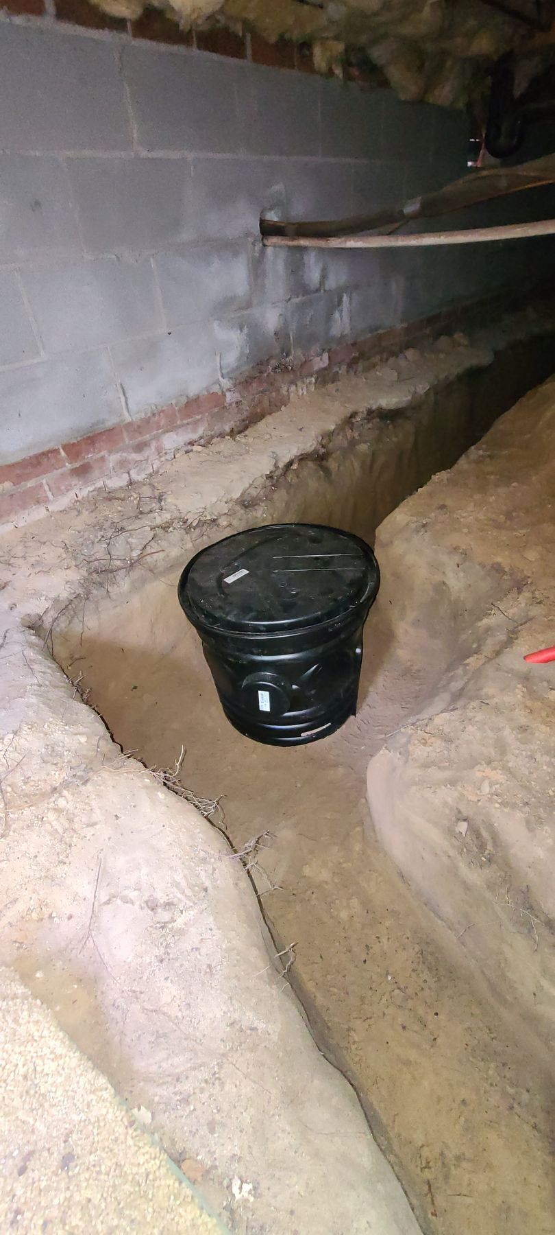 Black bucket in a narrow dirt trench beside a gray cinderblock wall inside a basement-like space