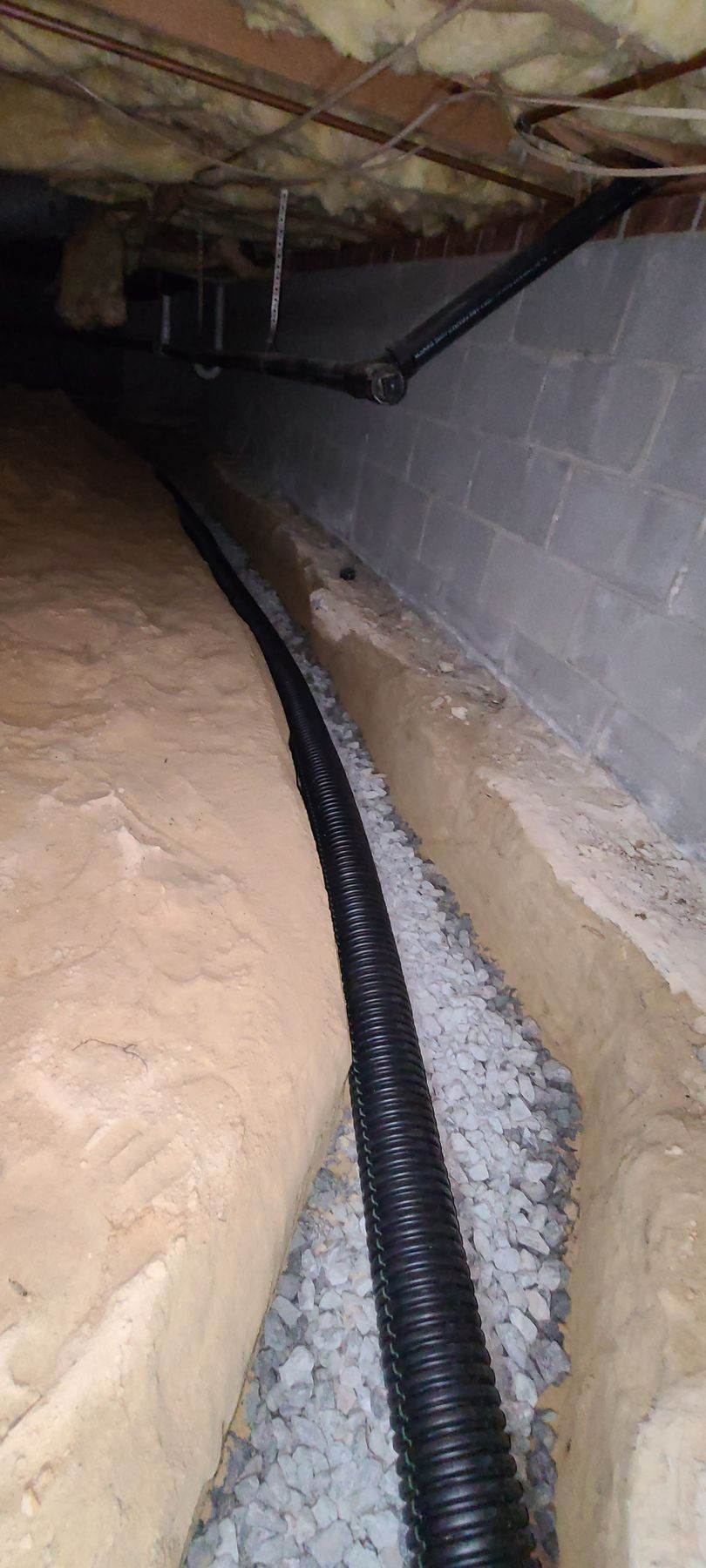 Basement crawlspace with a black corrugated pipe running beside a cinder block wall and exposed insulation.