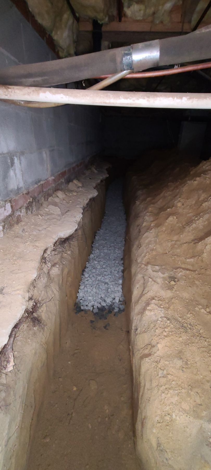 A narrow trench with a concrete pipe channel running through it under a low-ceilinged structure.