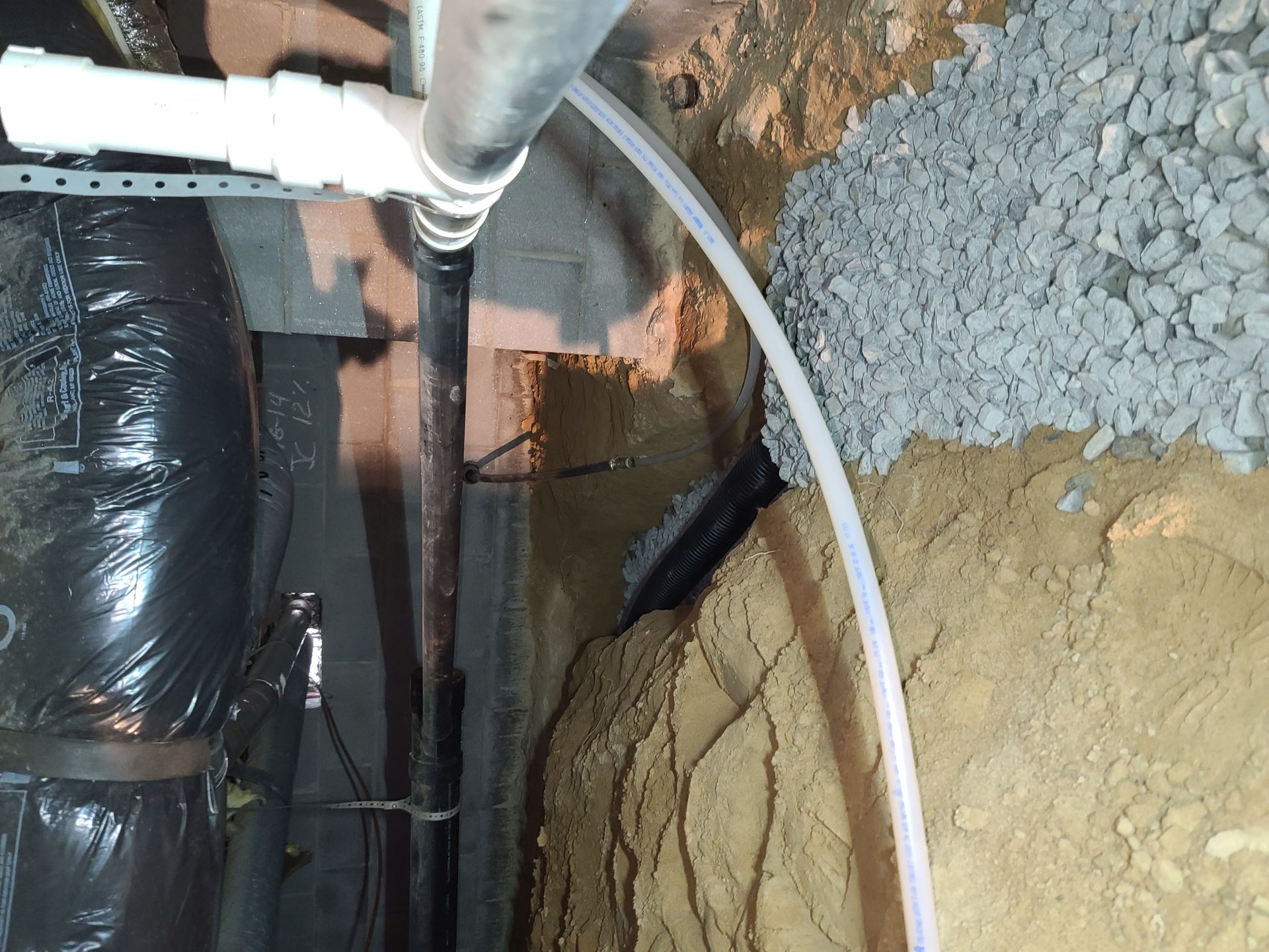 Insulation and pipes inside a wall cavity, with gravel and soil exposed around a foundation opening