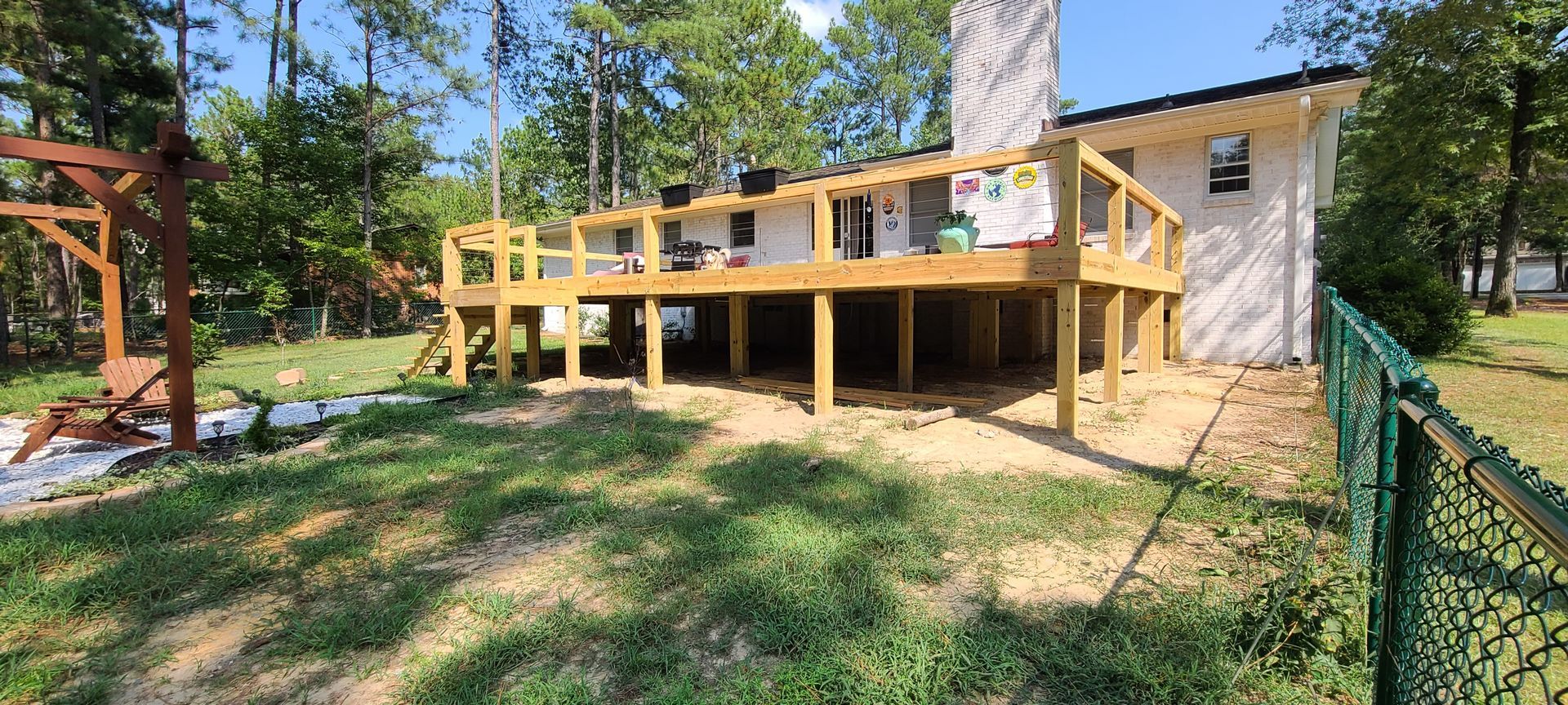 Wooden deck attached to a beige house in a sunny backyard with trees and a chain-link fence.