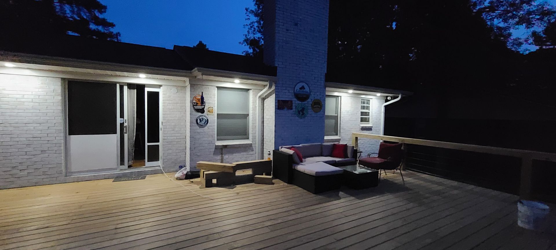 A lit wooden deck with patio furniture outside a white house at dusk.