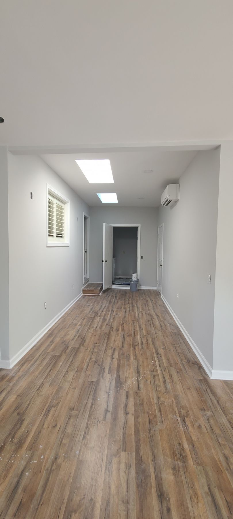 Empty narrow hallway with wood-look flooring, white walls, ceiling lights, and a doorway at the far end