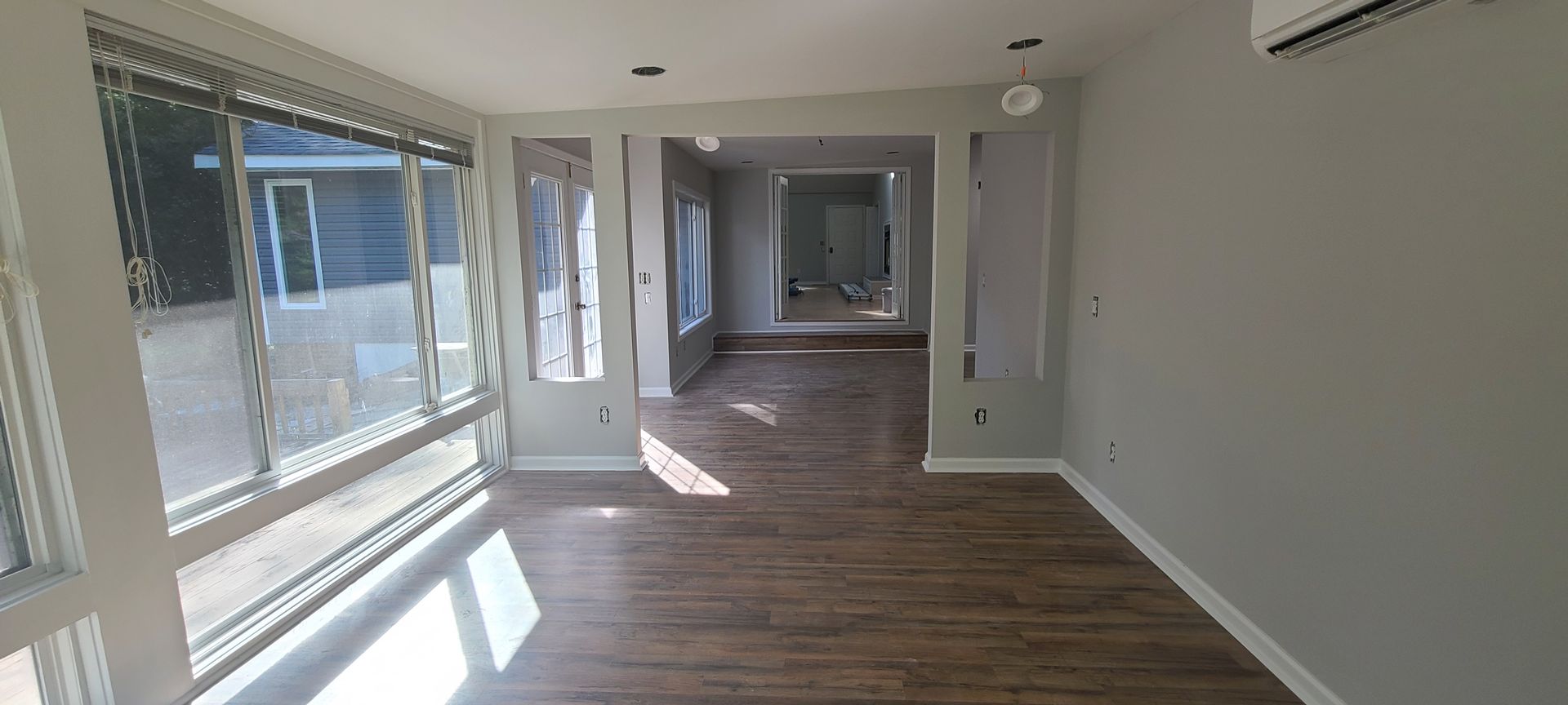 Empty modern room with wood flooring, white walls, large windows, and sunlight streaming in from the left