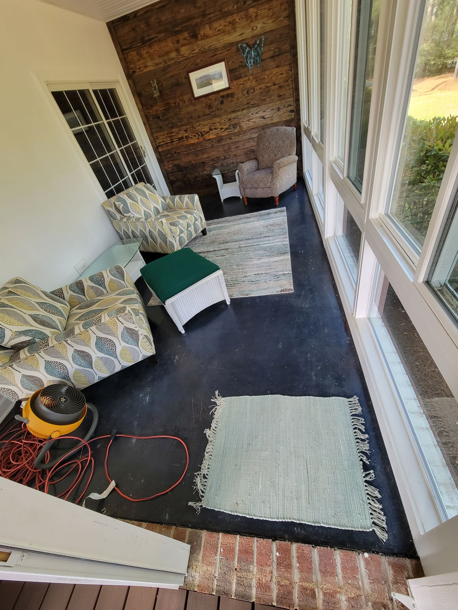 Small enclosed porch with patterned sofa, rugs, chair, and coiled hose beside windows.