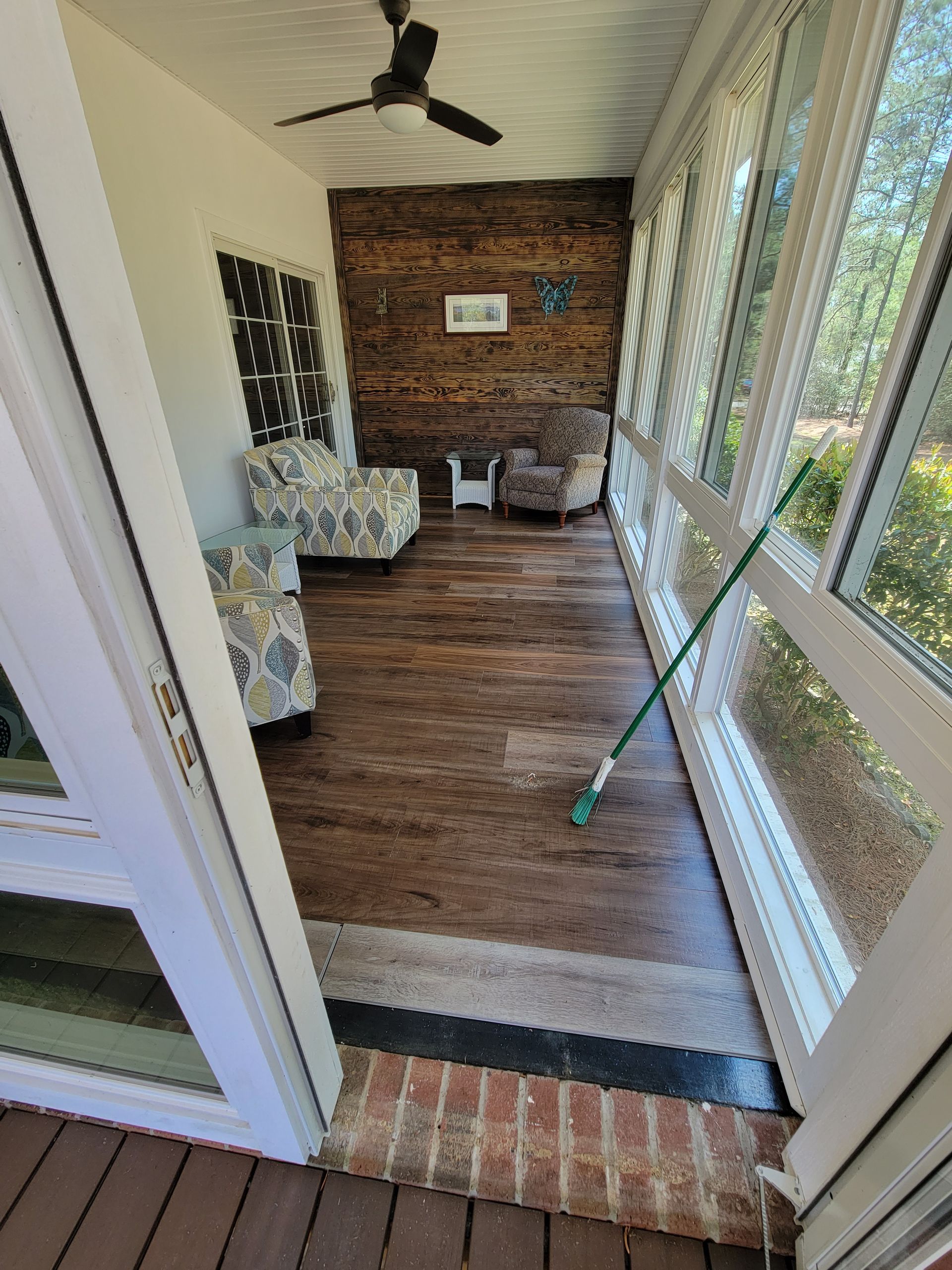 Sunroom with brick accent wall, wood floor, wicker chair, windows, and broom by the glass door.