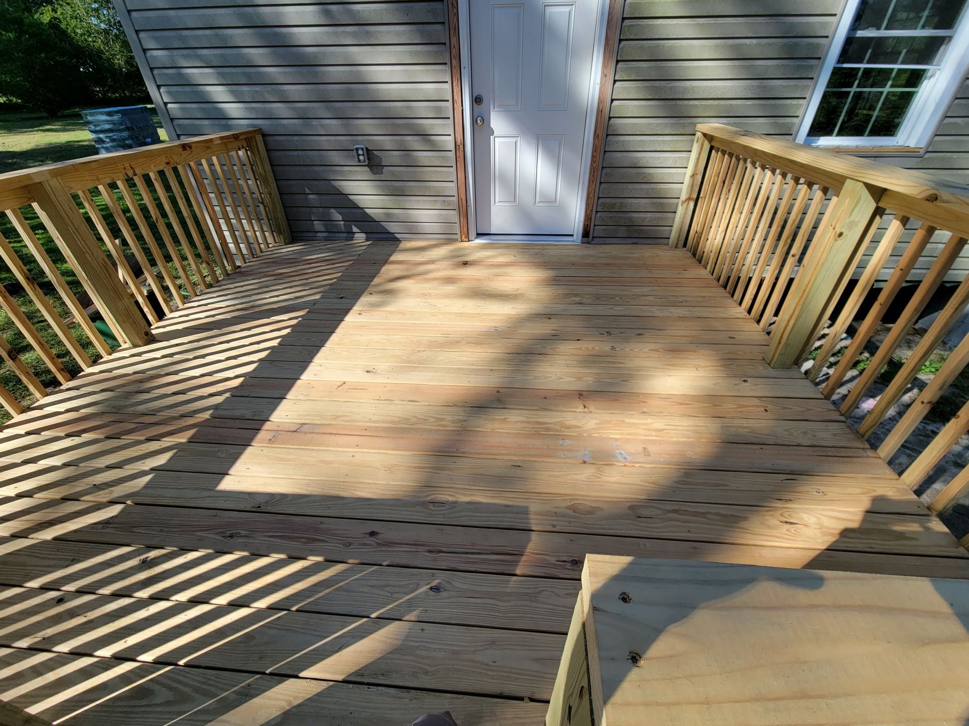 Wooden deck with railings leading to a white door on a house, lit by sunlight and shadows