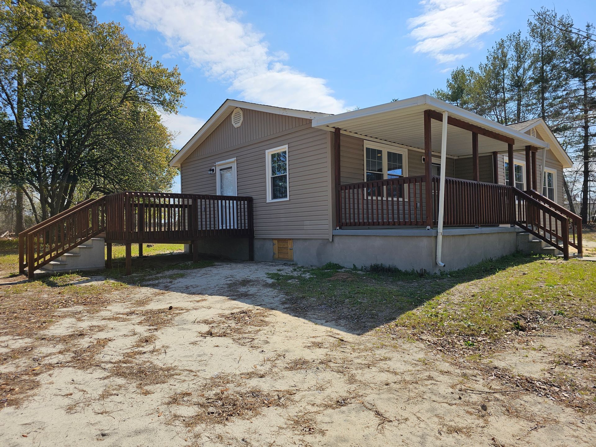 Single-story tan house with covered porch and wooden ramps, surrounded by trees and dirt yard