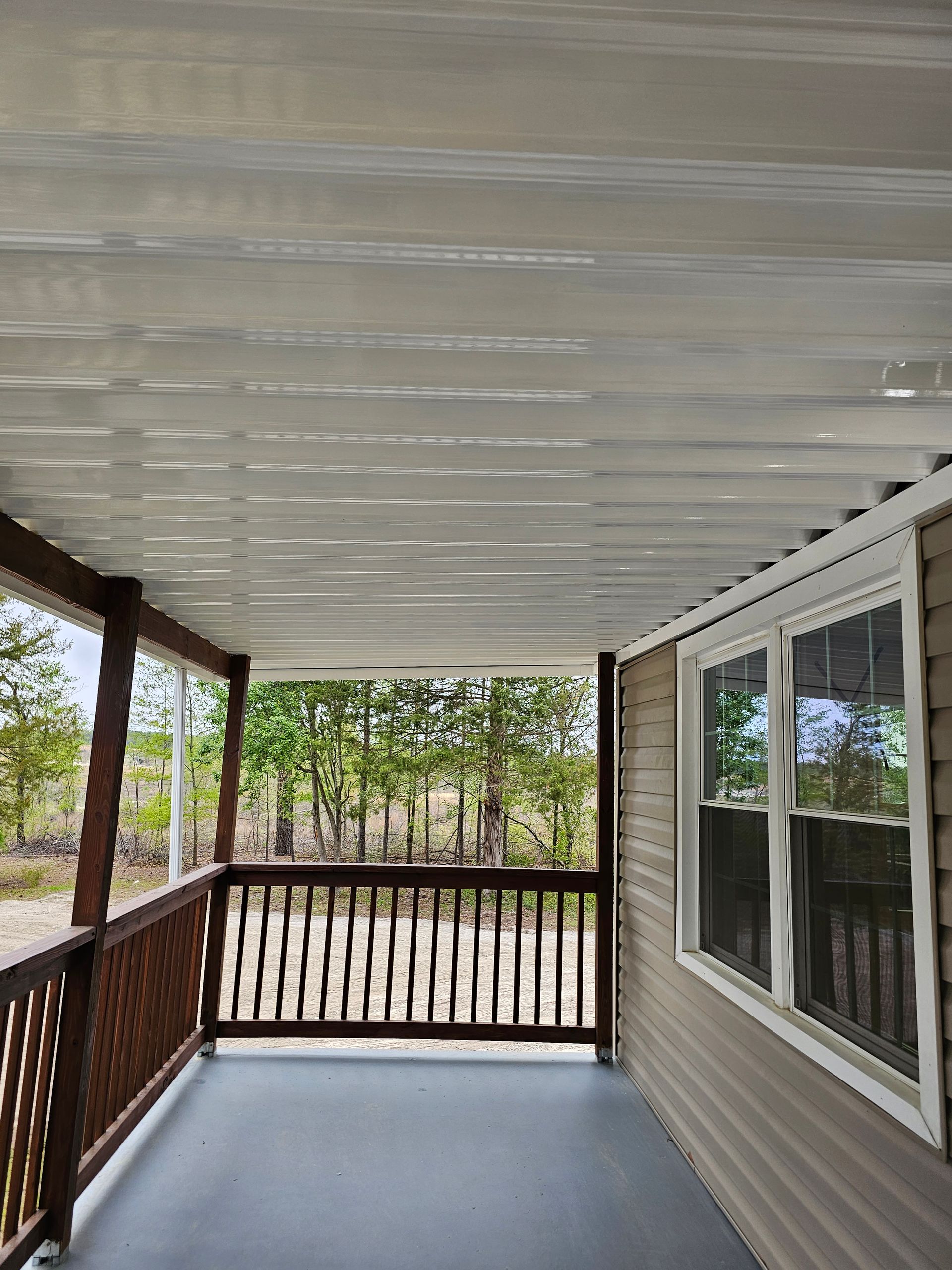 Covered porch with white ceiling, gray floor, wood railing, and windows overlooking trees