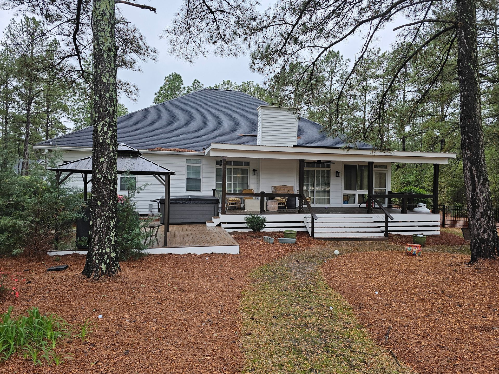 Single-story house with dark roof and porch, surrounded by pine trees and a leaf-covered yard