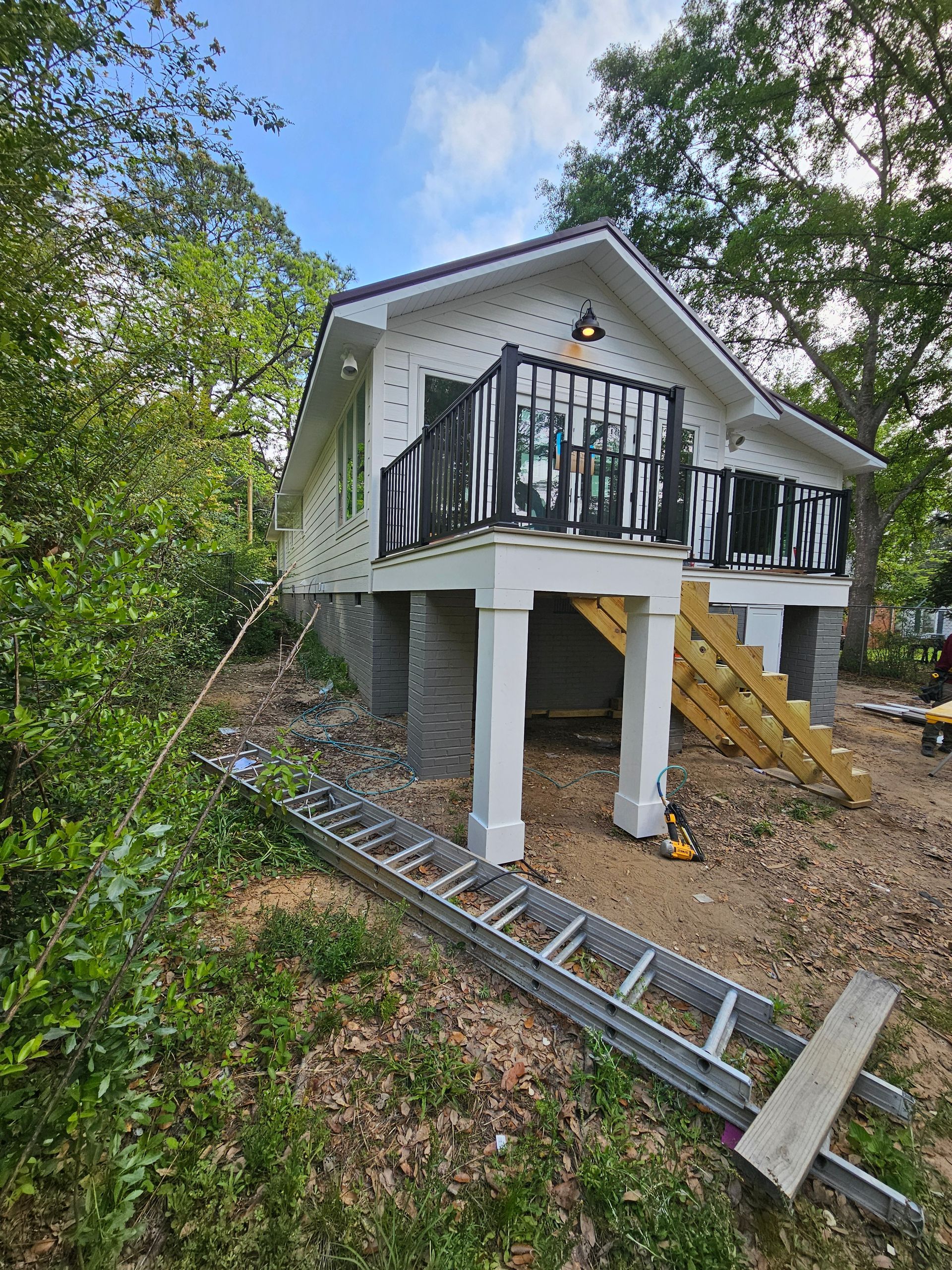 Small white house on stilts with black balcony railing, surrounded by trees and a ladder on the ground