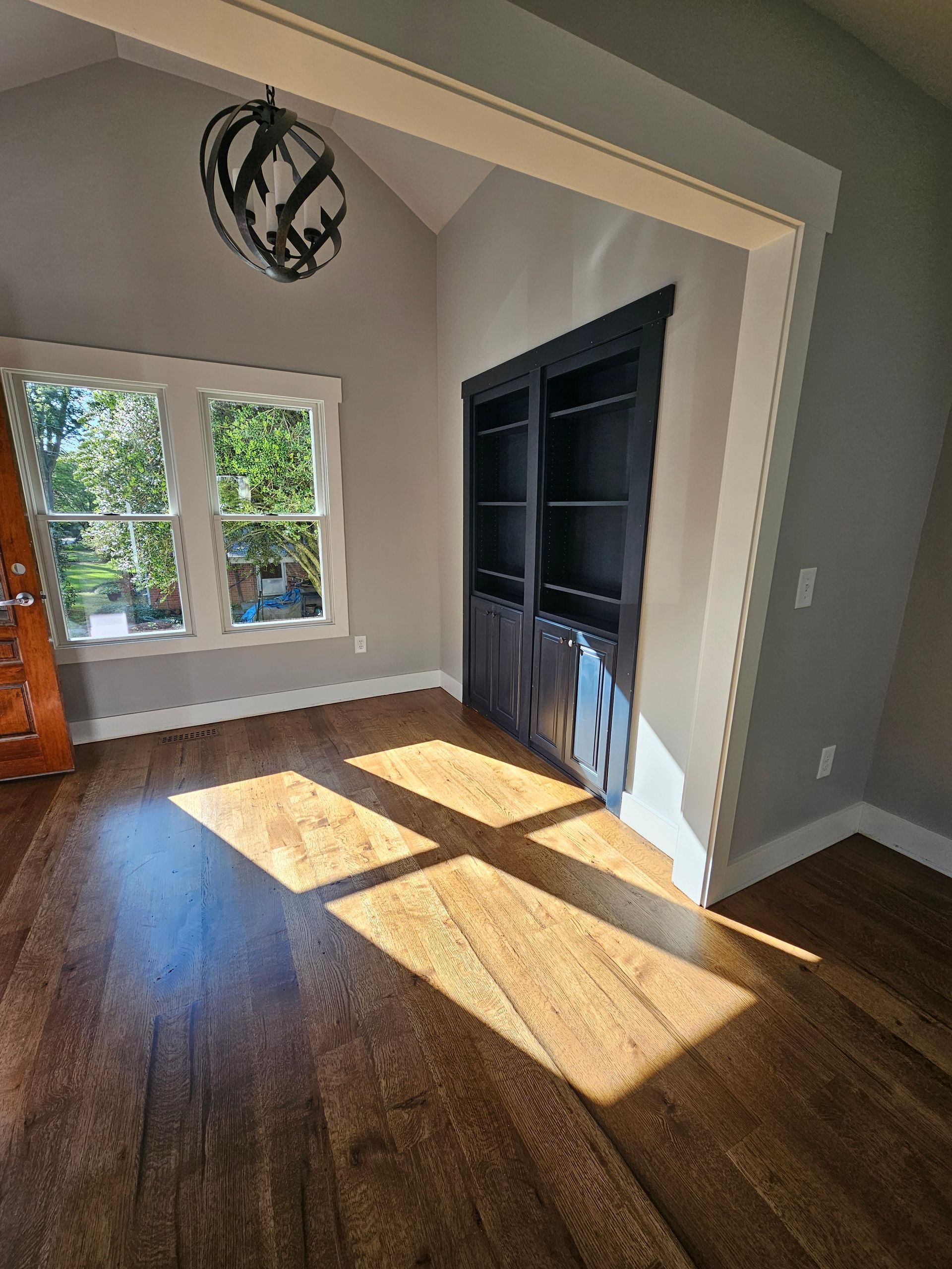 Bright empty room with wood floors, large window, and dark built-in shelving in sunlight.