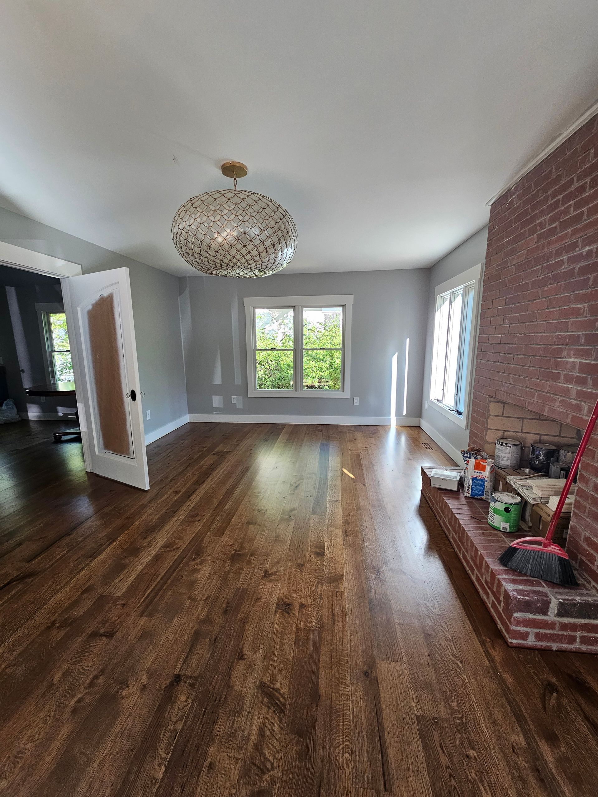 Empty living room with hardwood floors, brick fireplace, chandelier, and large windows letting in daylight