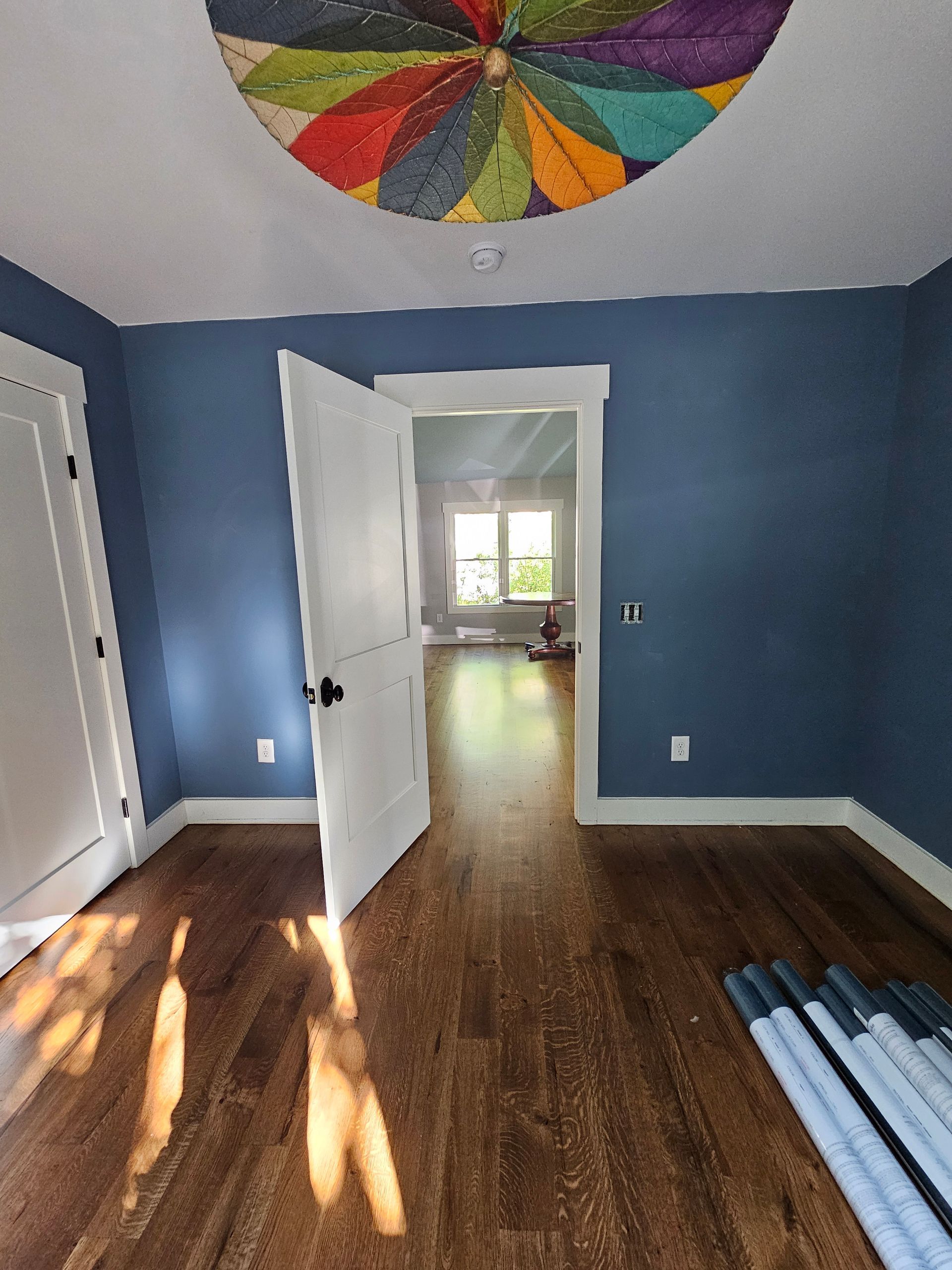 Empty blue bedroom with open white doors, hardwood floor, sunlight, and colorful ceiling fan decor.