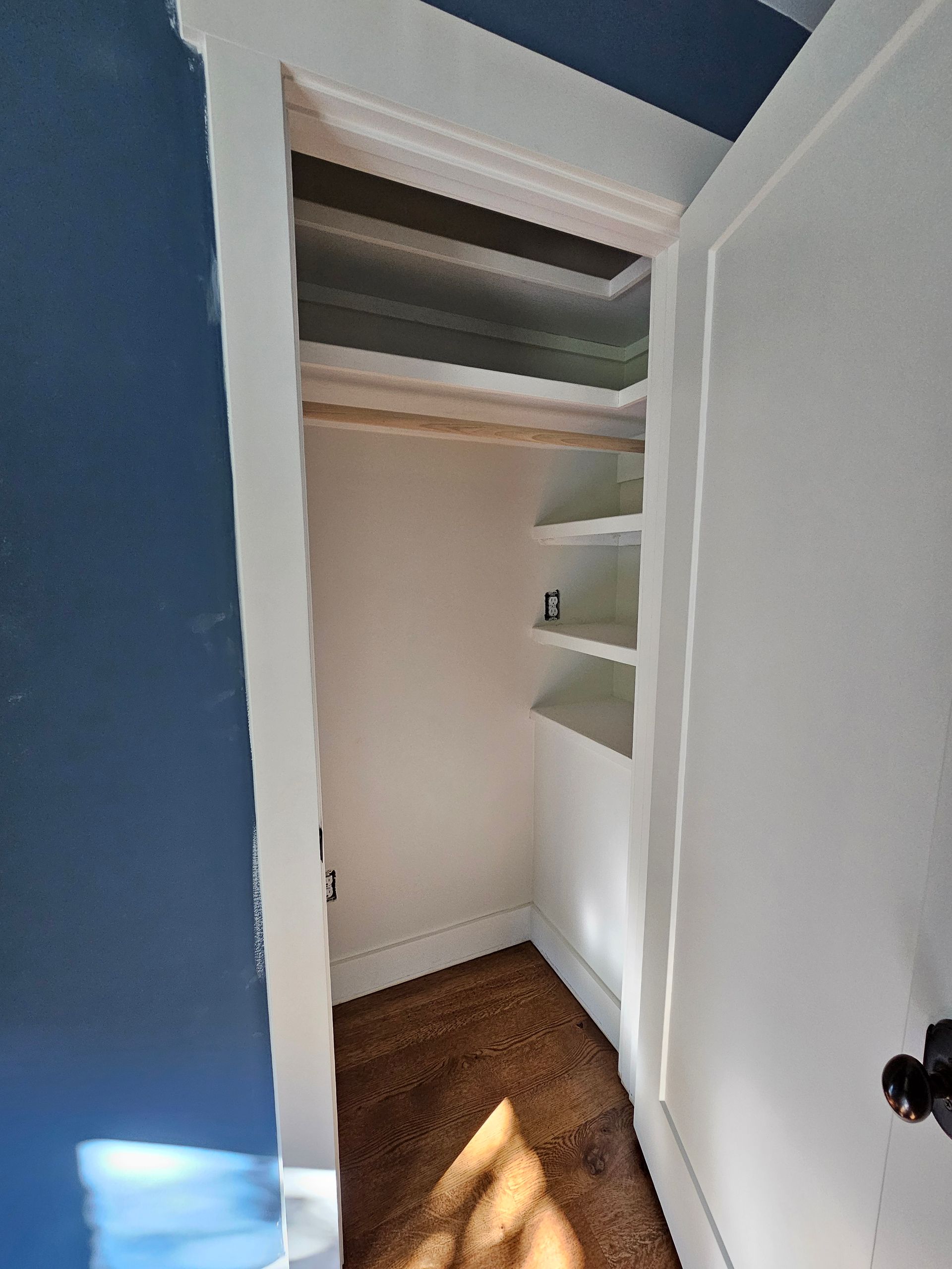 Open white closet with shelves and hanging rod in a blue-walled room, sunlight on wood floor