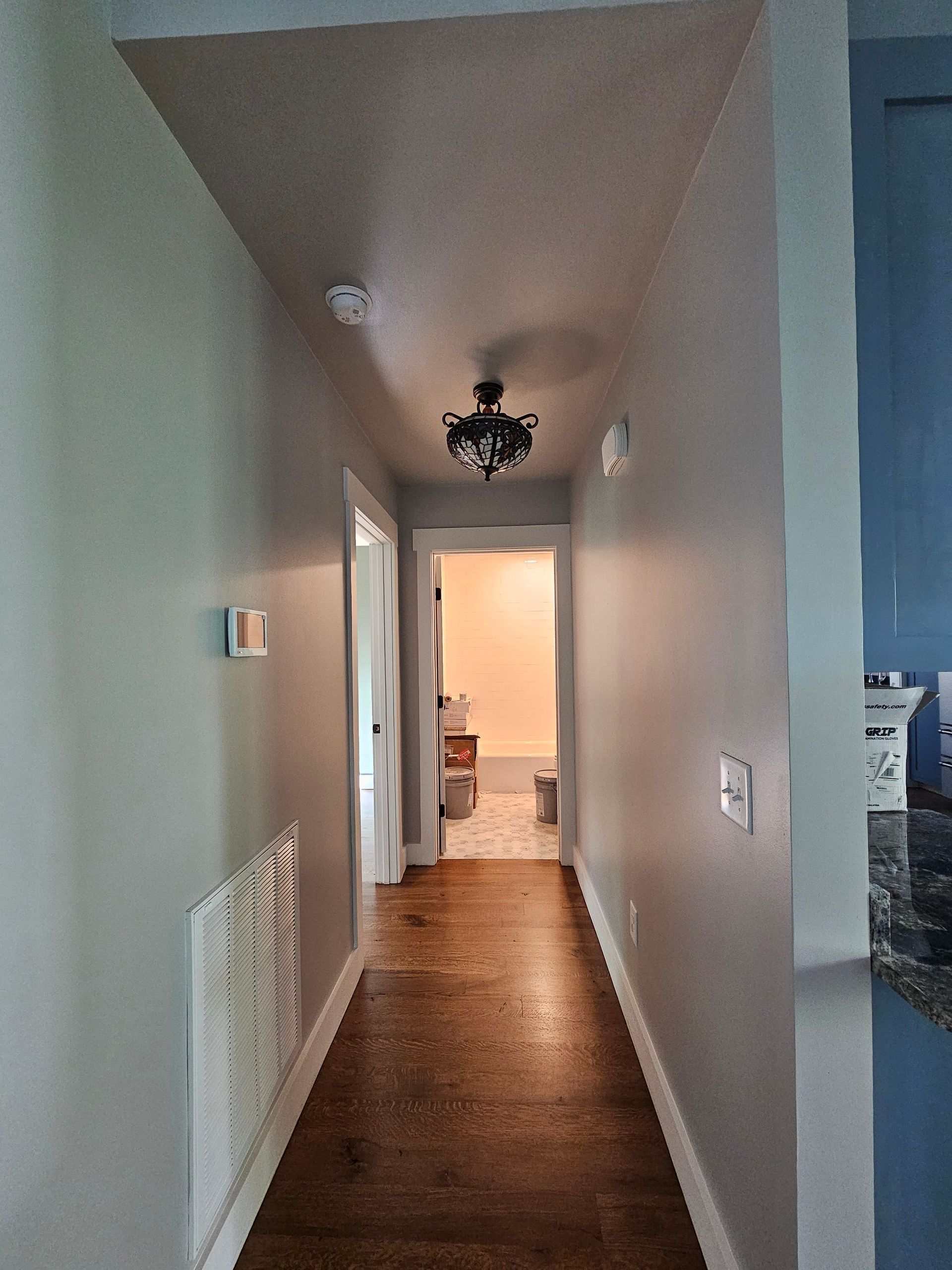 Narrow hallway with gray walls, wood floor, and ceiling lights leading to a bright room at the end