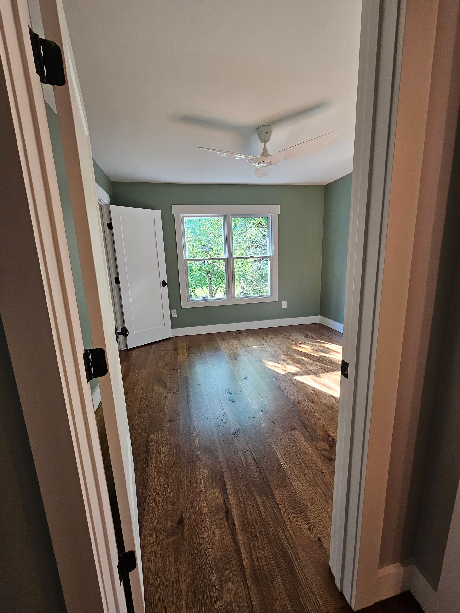 Empty bedroom with green walls, hardwood floor, and a window letting in daylight.