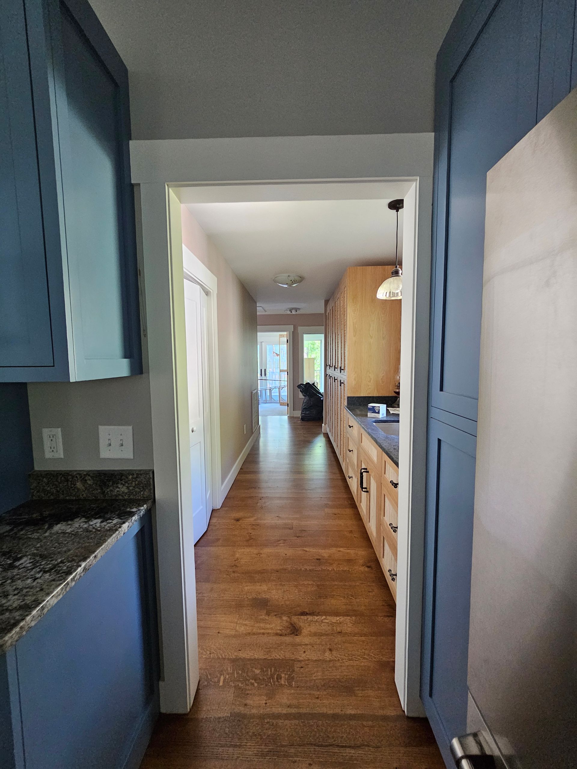 Narrow kitchen hallway with blue cabinets, wood floor, and a doorway leading to a bright room.