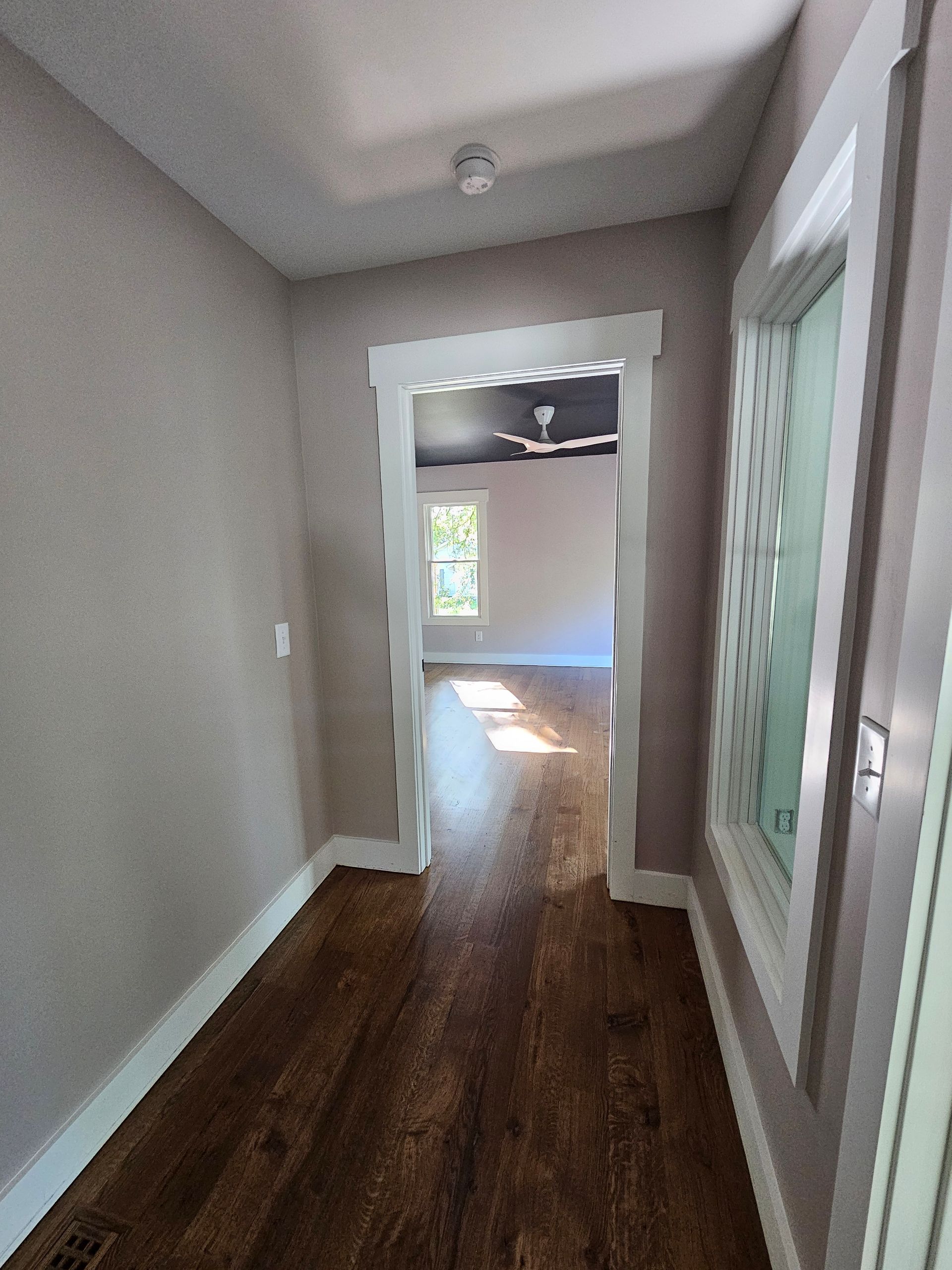 Empty hallway with gray walls, dark wood floor, and sunlight from a bright room ahead
