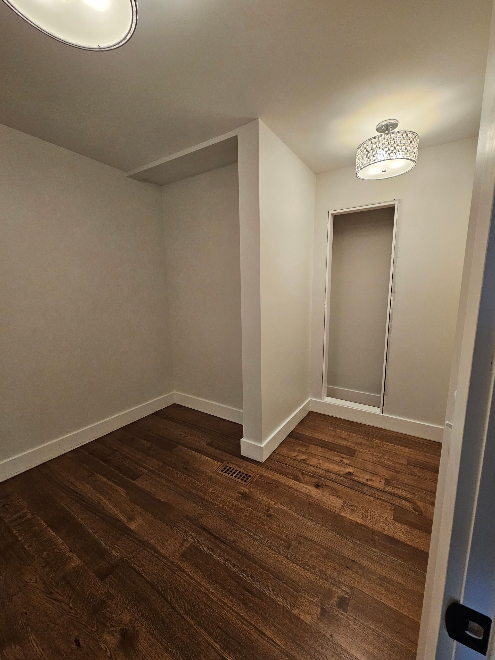 Empty room with wood floors, beige walls, and a ceiling light, viewed toward a hallway opening.