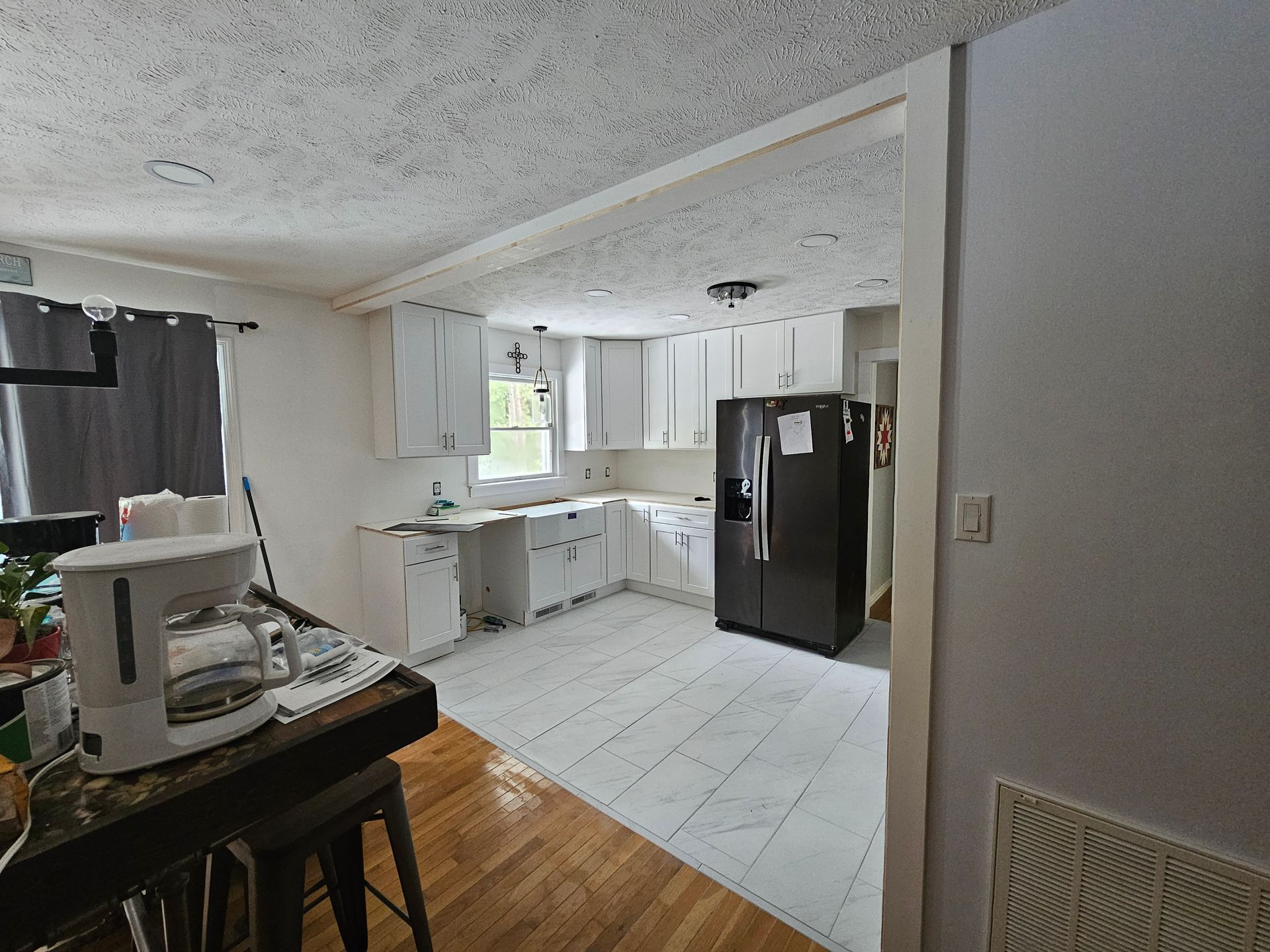 Kitchen with white cabinets, black refrigerator, and tiled floor opening into a wood-floored room.