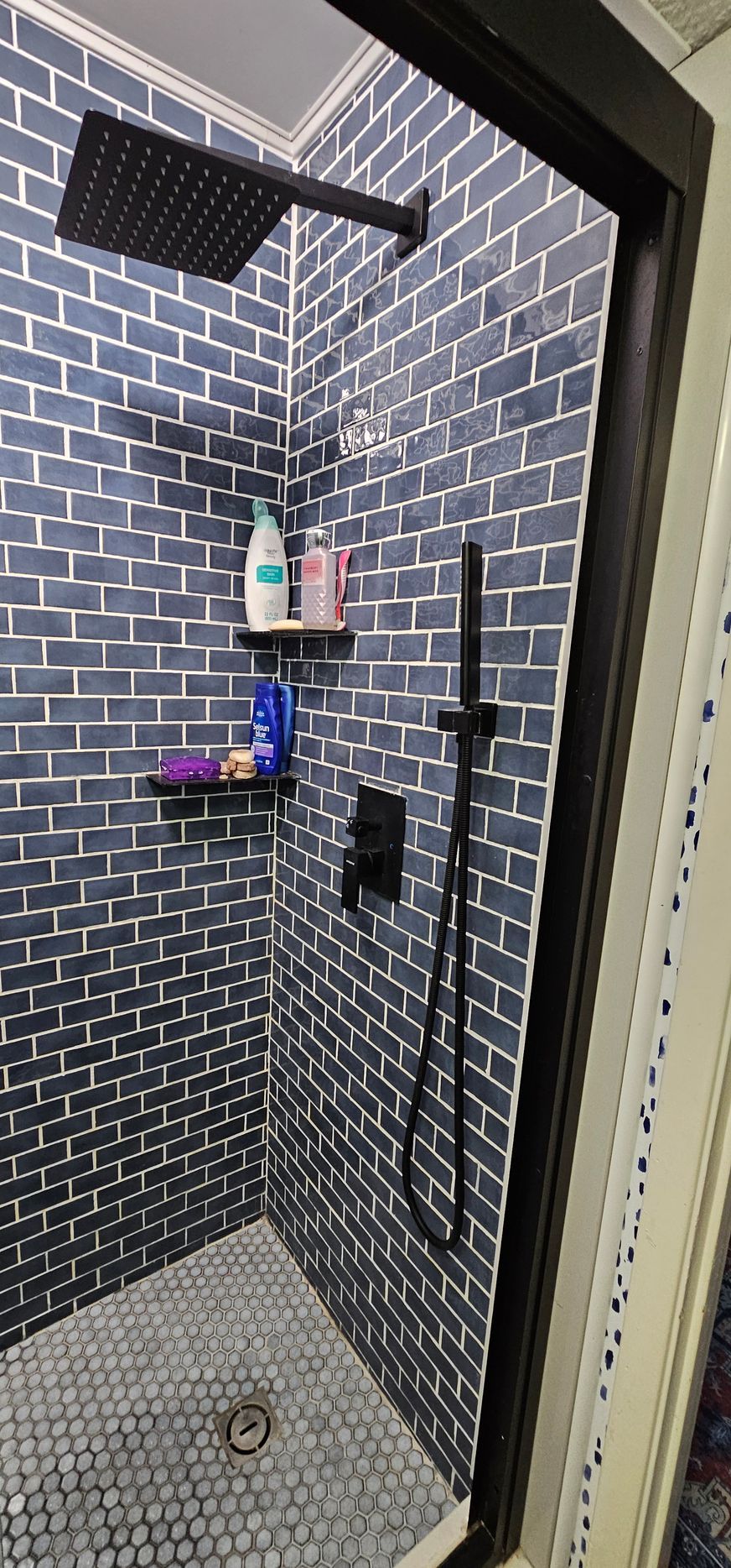 Black-tiled shower with rain showerhead, handheld sprayer, and wall-mounted toiletries.