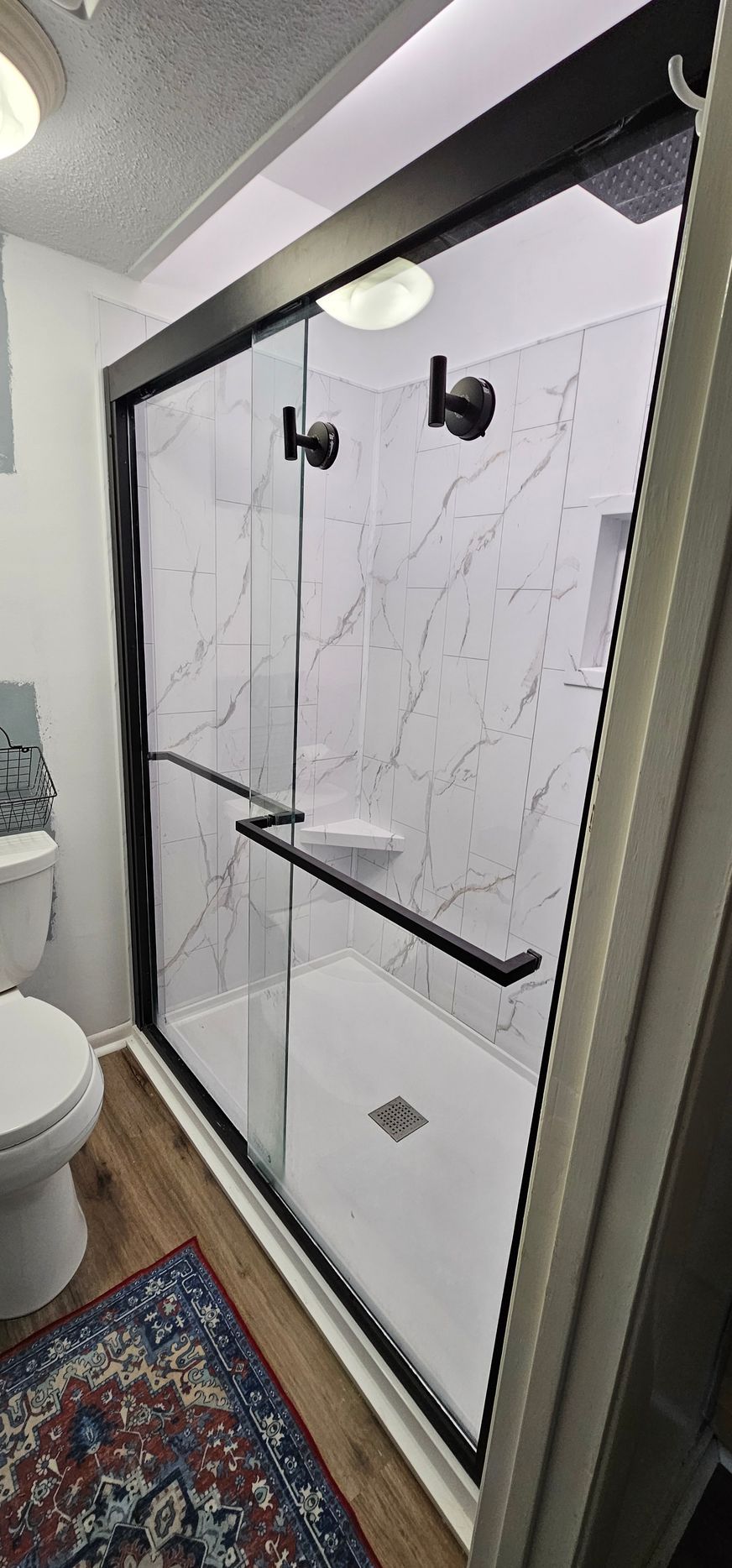 Bathroom shower with clear glass doors, marble-look tile, and black fixtures in a narrow space