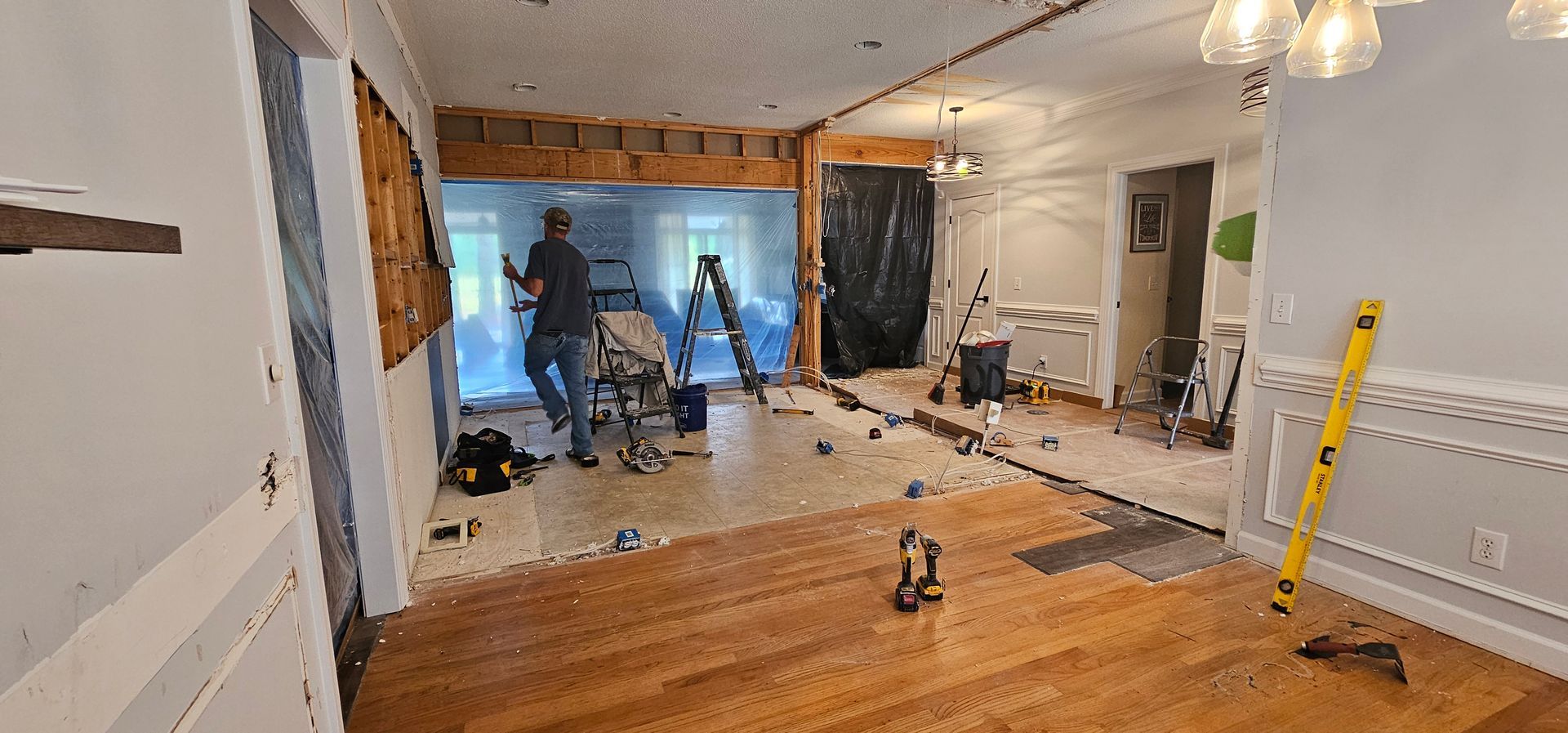 Renovated living room with hardwood floors, workers, tools, and daylight through a large window.