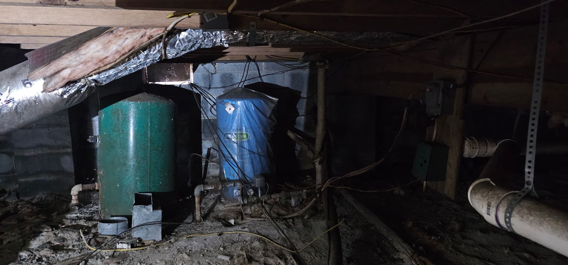 Basement crawlspace with two large green and blue water tanks amid pipes and debris