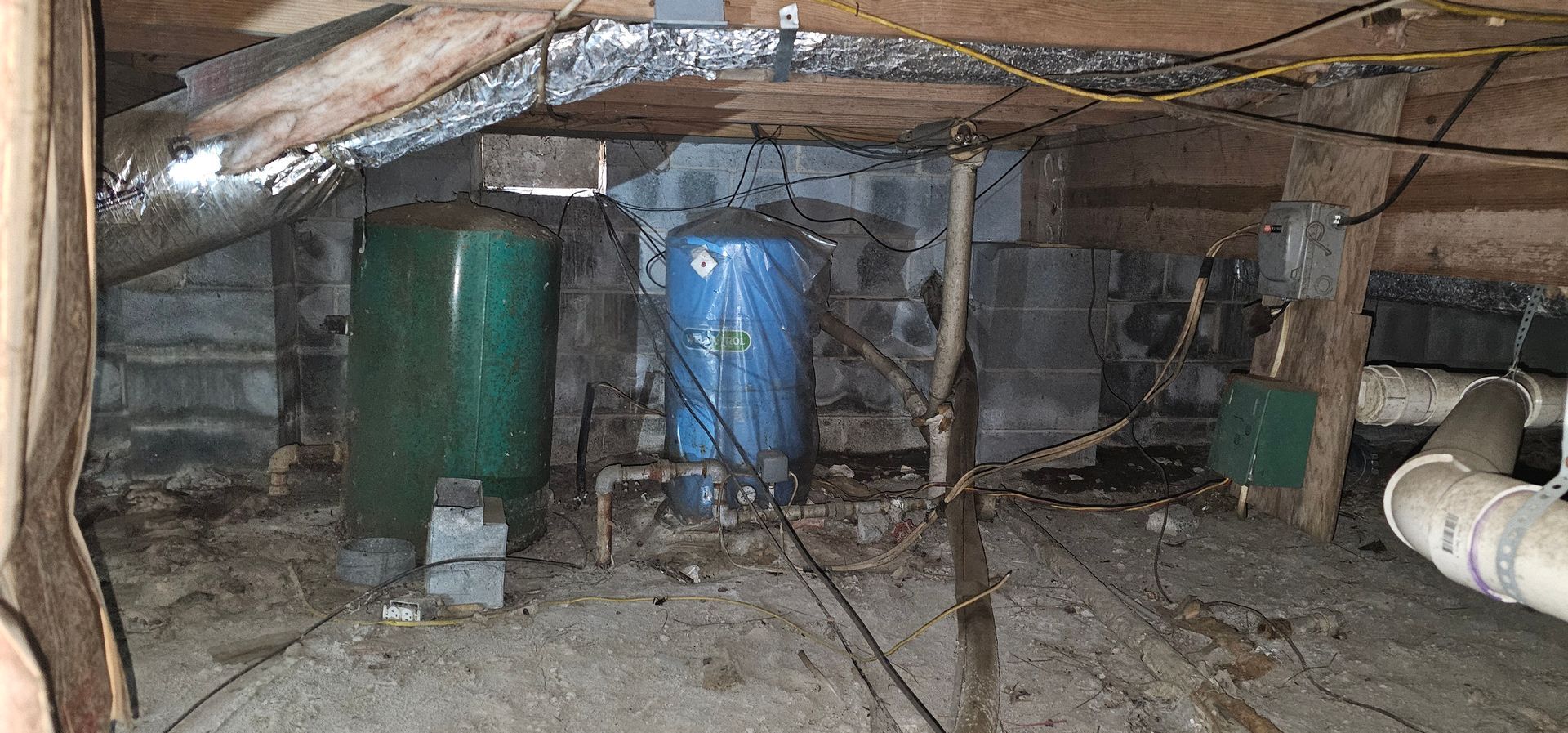 Under-floor crawlspace with two blue and green tanks among pipes and debris