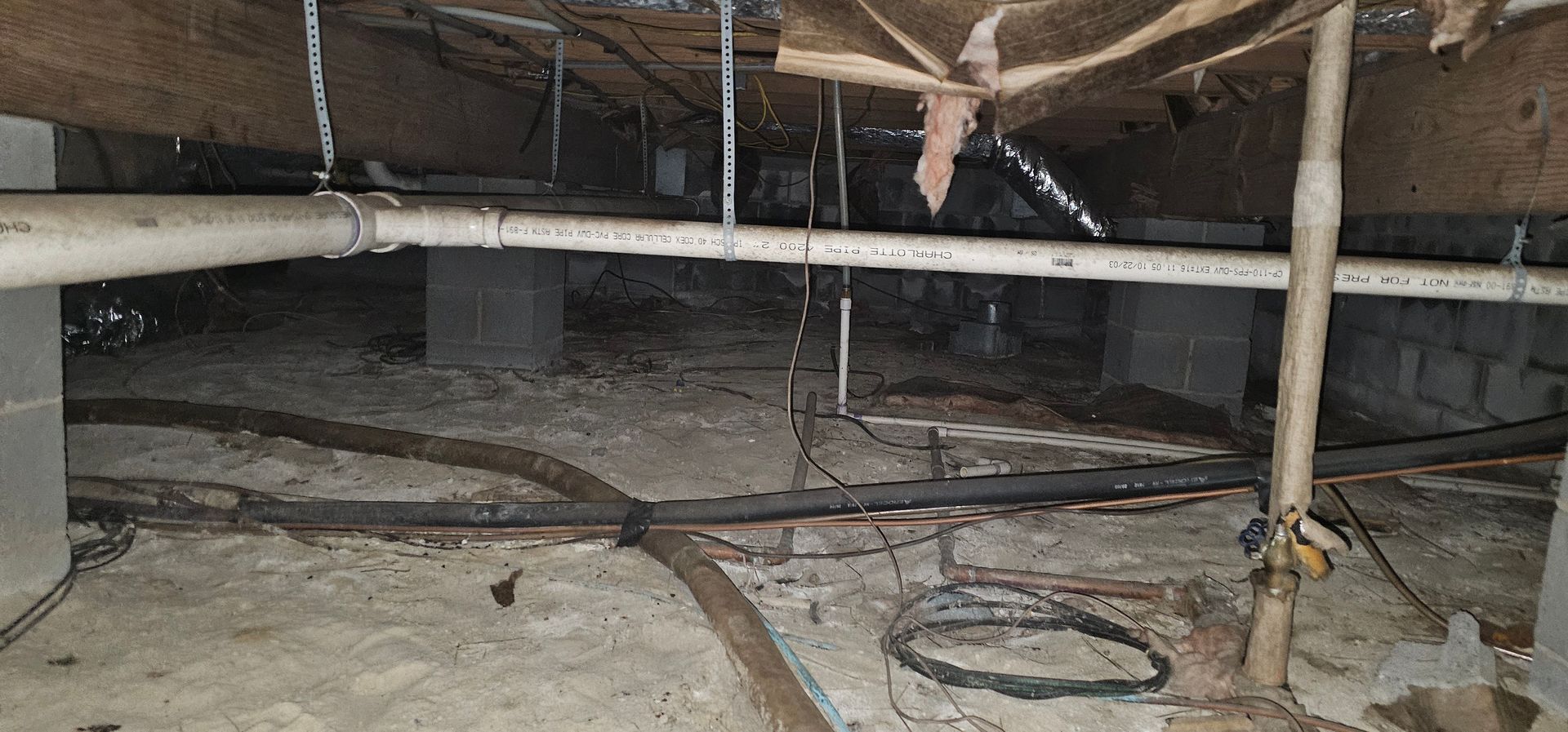 Crawl space under a house with exposed pipes, beams, and scattered cables on a dirt floor