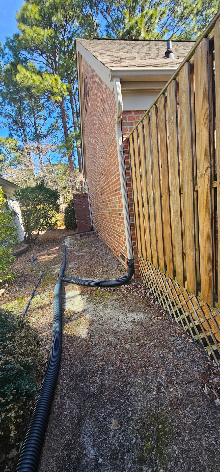 Narrow gravel side yard beside a brick house and wooden fence under blue sky