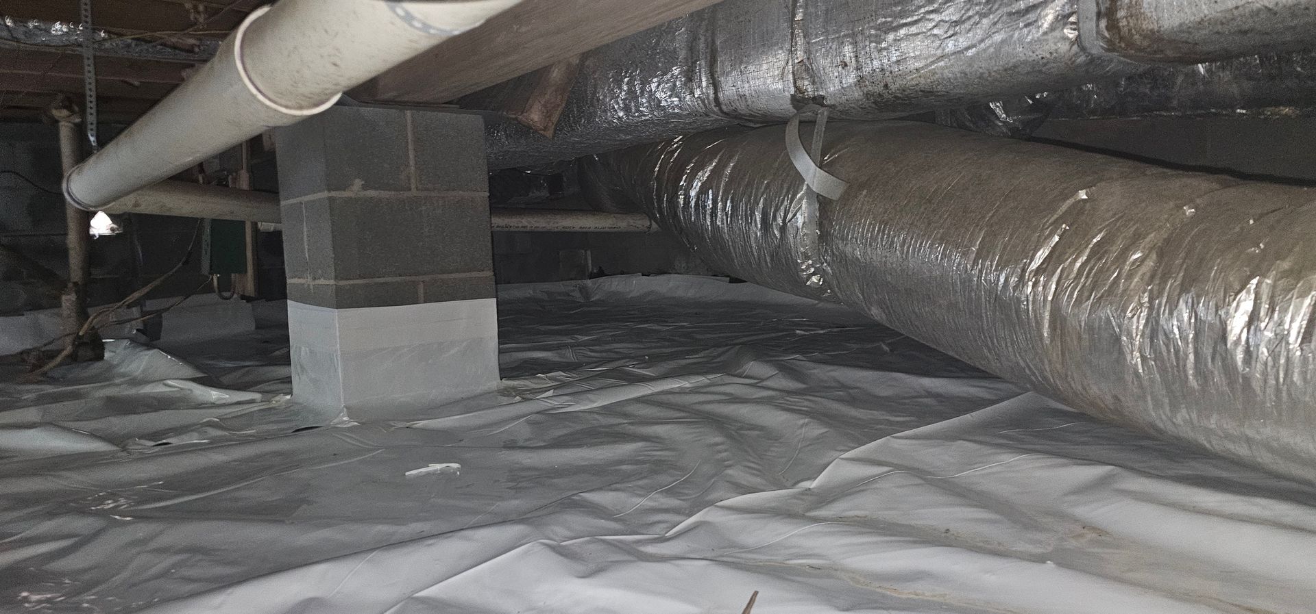 Crawlspace with insulated ductwork and pipes above a white plastic vapor barrier