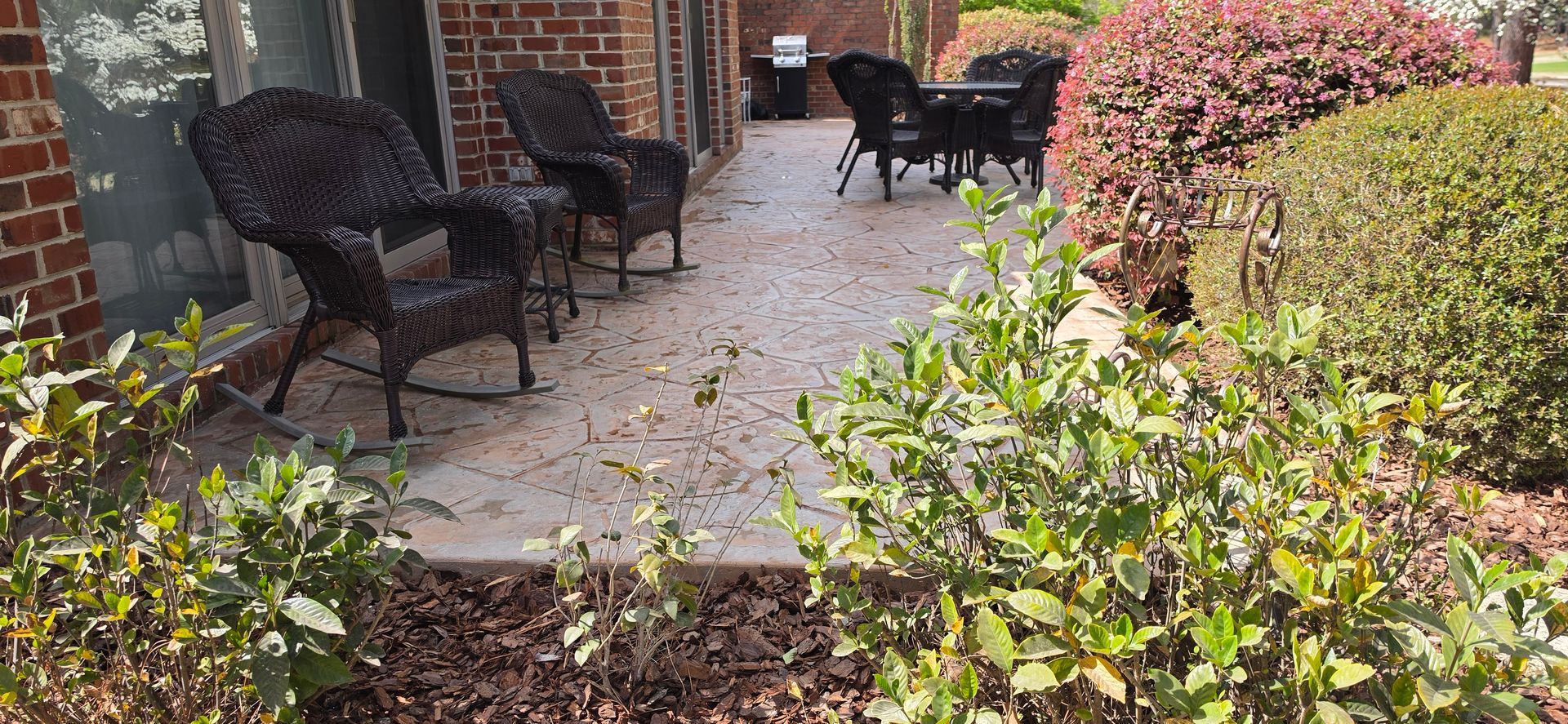Brick patio with black wicker chairs, potted plants, and shrubs beside a house