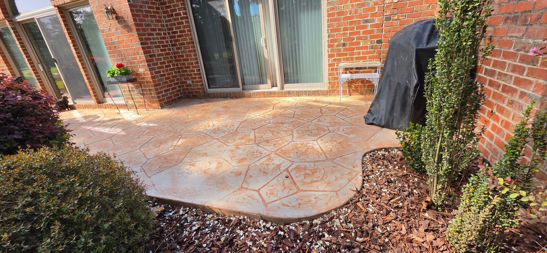 Brick patio outside a house with sliding glass doors, potted greenery, and a grill covered by a black cover