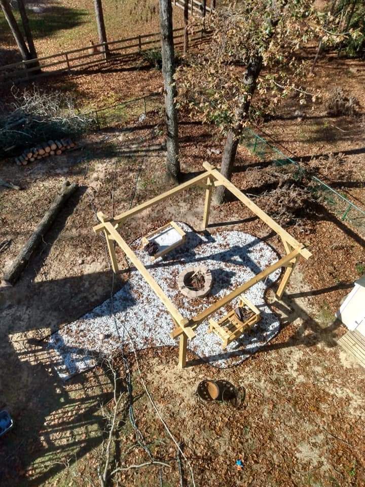 Yellow square frame over a fire pit in a wooded yard, with tools and tarp on the ground.