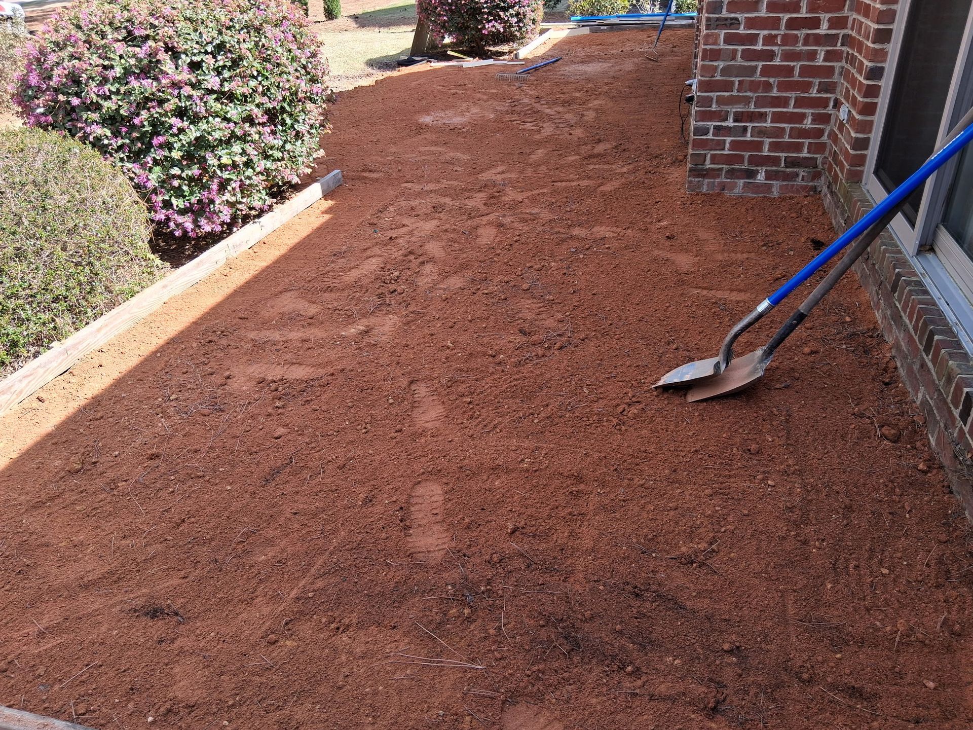 Garden bed with red mulch beside a brick wall, with a shovel leaning on the right side.