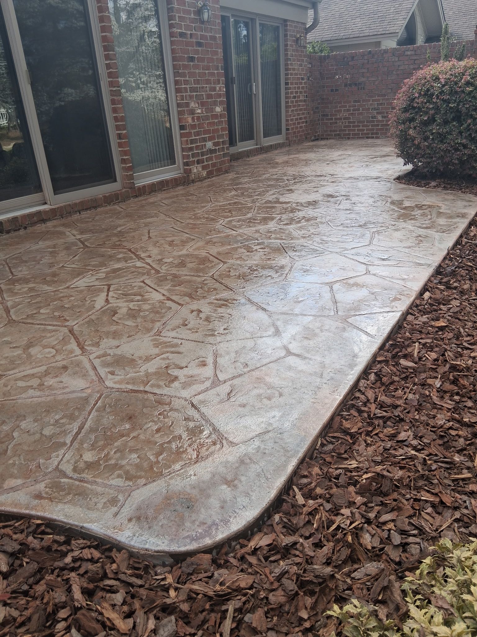 Rain-soaked stamped concrete patio beside a brick house and shrubs